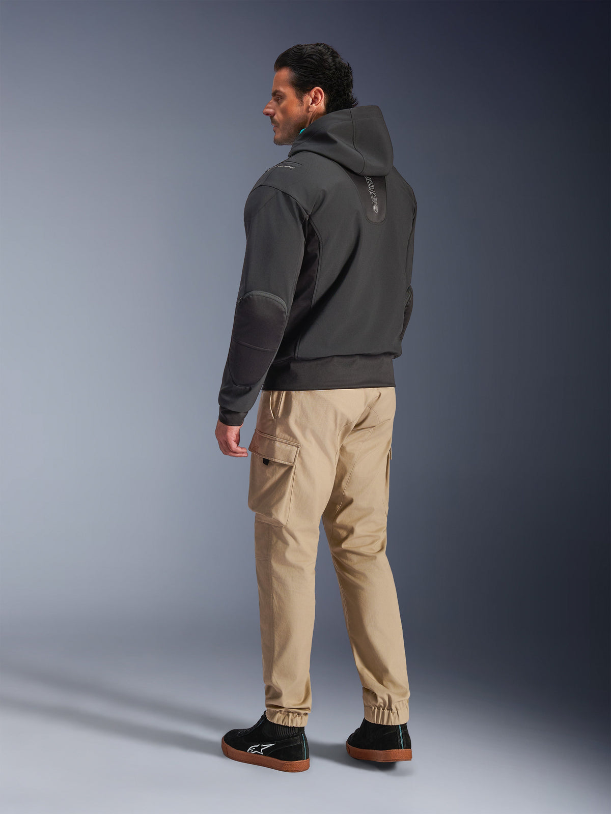 Alpinestars Moblast Wasserdichte Jacke, schwarzer Motorrad-Tech-Hoodie, Seiten- und Rückansicht, getragen mit beigefarbener Cargohose und schwarzen Sneakern, Kapuzendesign, technische Textilkonstruktion, moderne urbane Ästhetik.
