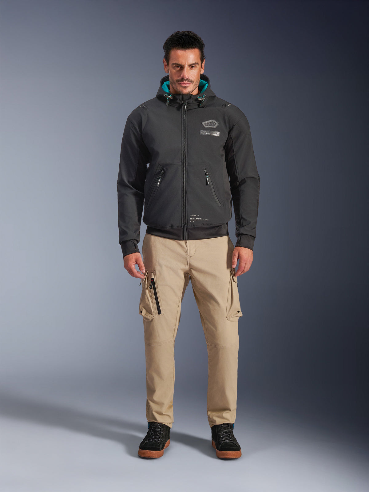Alpinestars Moblast wasserdichte Jacke, schwarzer urbaner Motorrad-Hoodie mit teal-farbenem Kapuzeninnenfutter, getragen mit sandfarbenen Cargo-Hosen und schwarzen High-Top-Stiefeln, moderne Ganzjahres-Fahrausrüstung