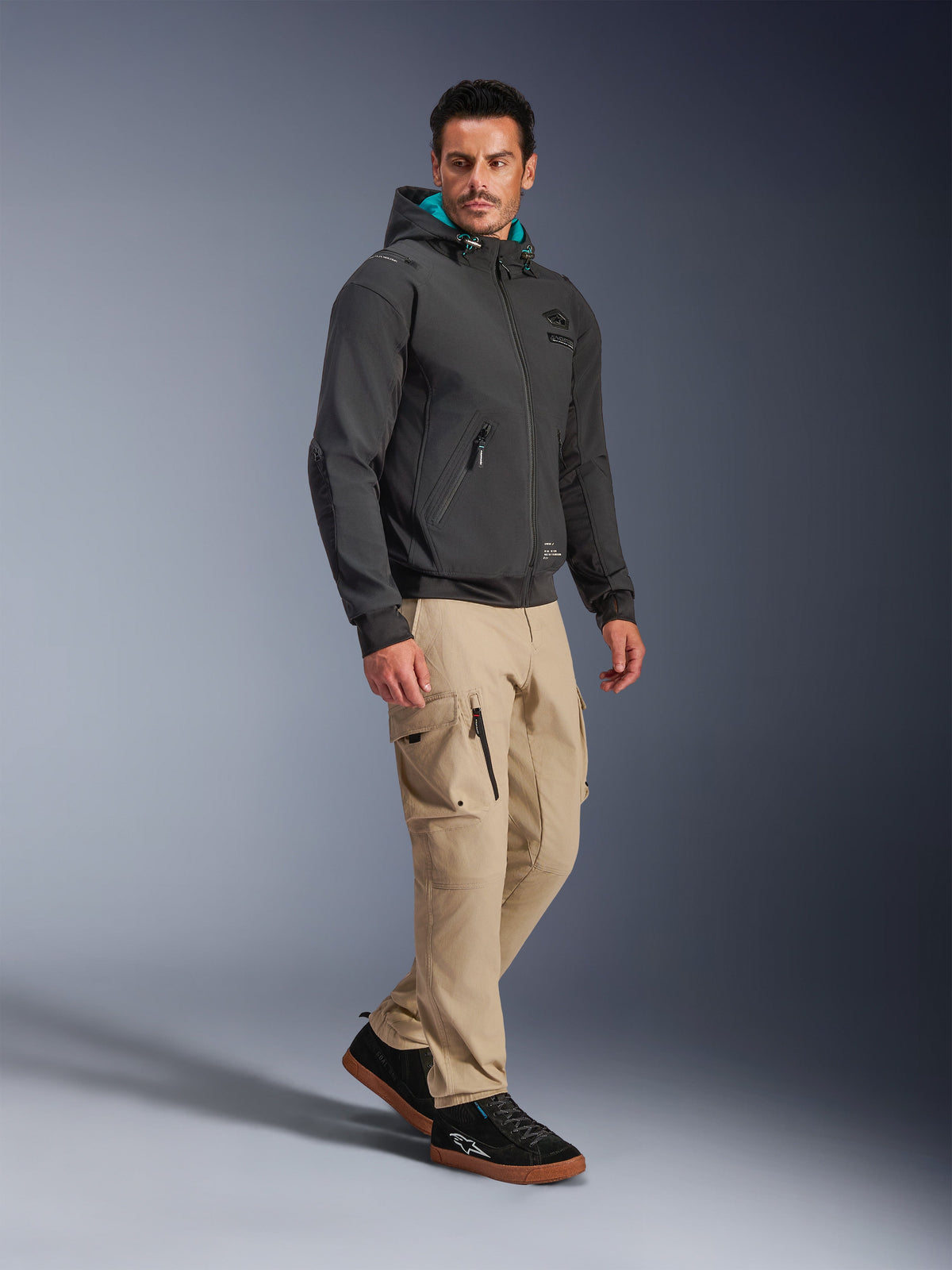 Männliches Model trägt die Alpinestars Moblast Waterproof Jacke in Schwarz, urbaner Motorrad-Hoodie mit türkis gefütterter Kapuze, kombiniert mit khakifarbener Cargohose und schwarzen Sneakern, modernes minimalistisches Design mit technischem Stretchgewebe für das Pendeln bei jedem Wetter