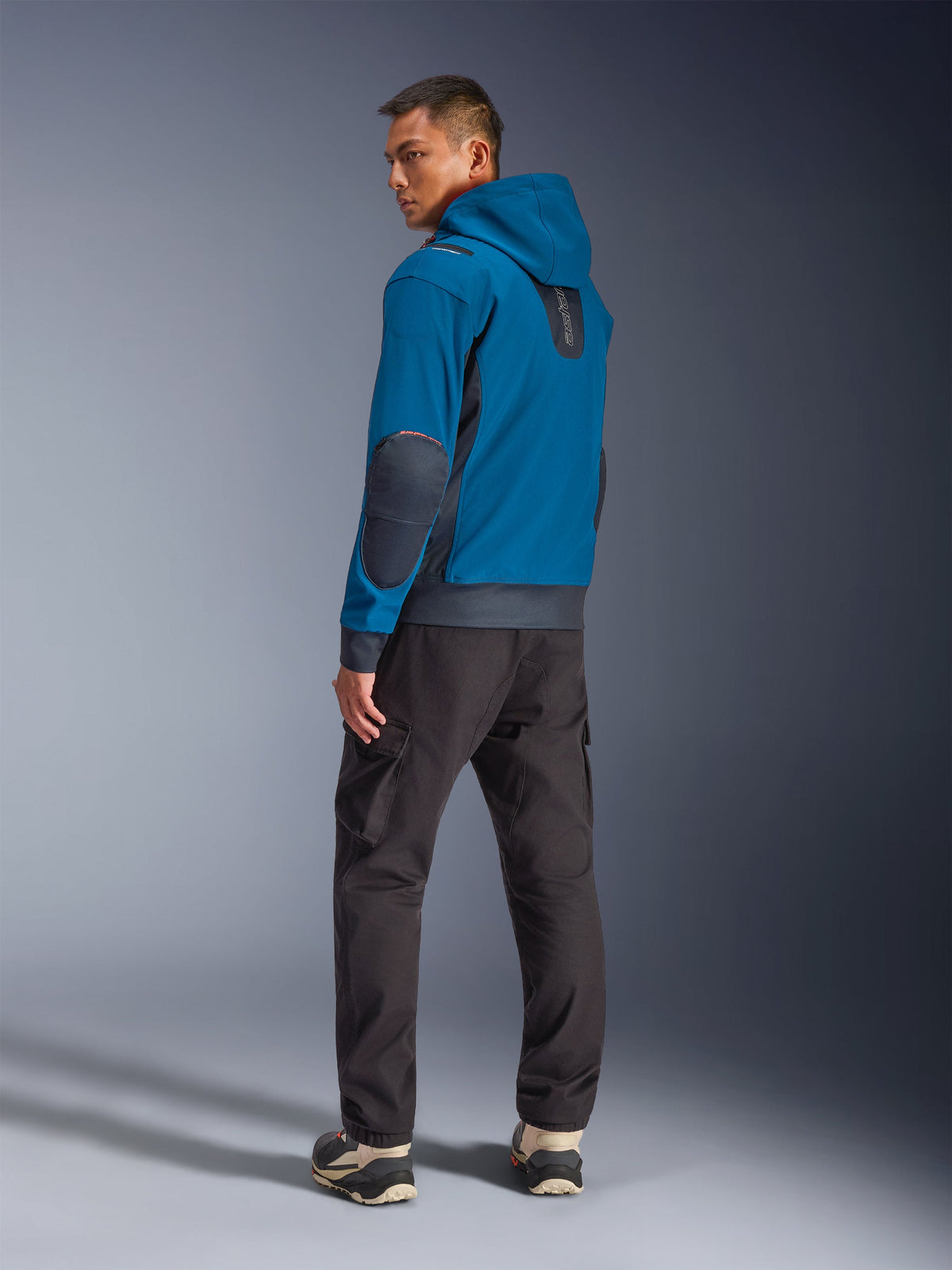 Alpinestars Moblast WP Jacke in Dark Petrol und Dark Navy, Rückansicht am Model mit Kapuzendesign, dunkelblauen Ellbogen-Patches und Seiteneinsätzen, kombiniert mit einer dunklen Cargohose und technischen Sneakern, moderne urbane Motorradbekleidung.