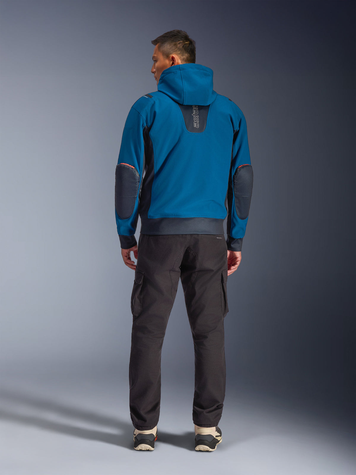 Alpinestars Moblast WP Jacke, urbane Motorradjacke, Dark Petrol und Dark Navy Blau, Rückansicht mit Kapuze und verstärkten Ellbogen-Patches, getragen mit dunkler Cargohose, elegante Allwetter-Ausrüstung für Pendler