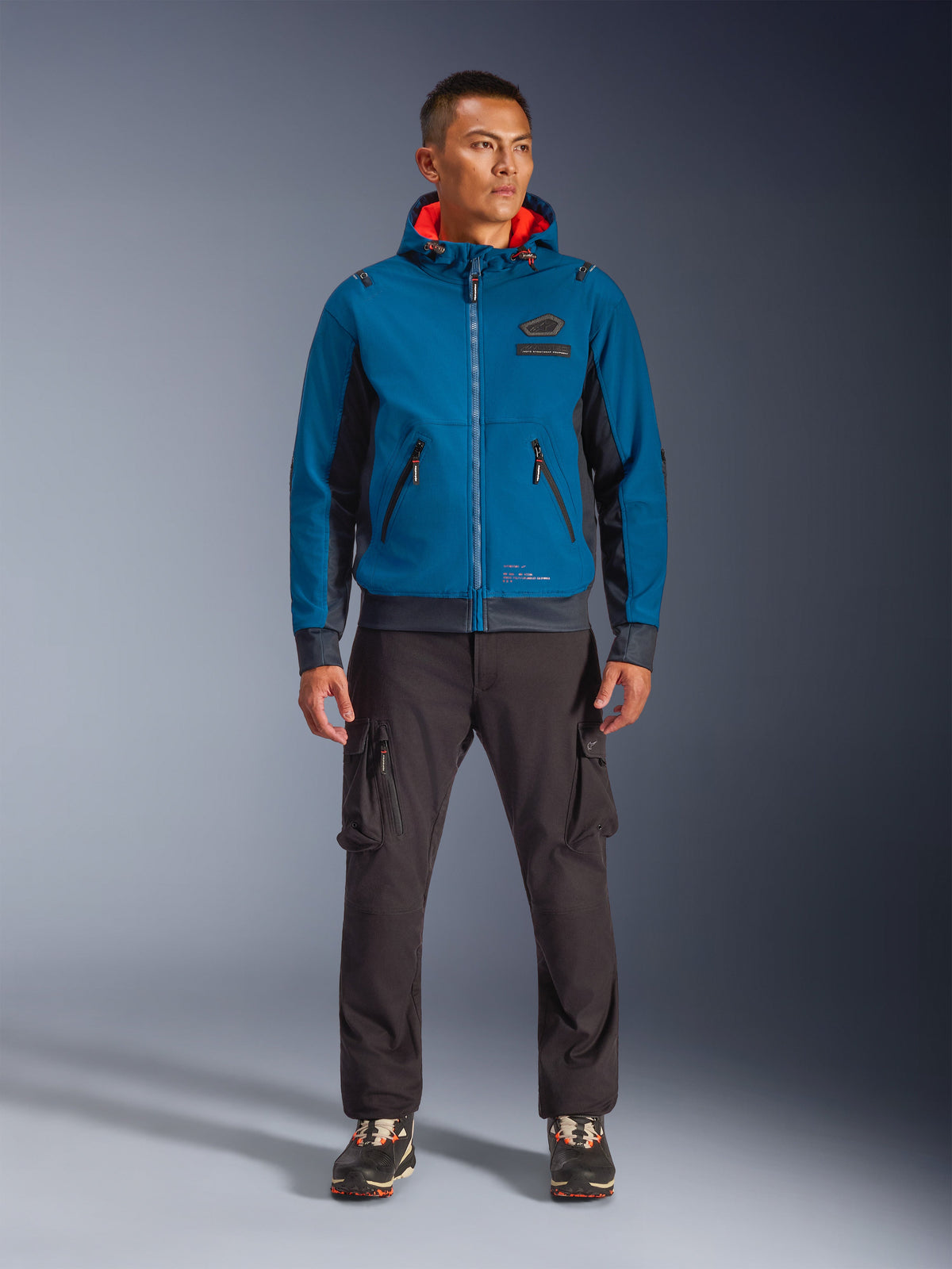 Alpinestars Moblast Waterproof Jacke, Motorradjacke mit Kapuze in Dunkelpetrol und Dunkelnavy mit orangem Innenfutter, getragen von einem männlichen Model mit dunkler Cargohose und schwarzen Sneakern, moderne urbane Fahrerausrüstung