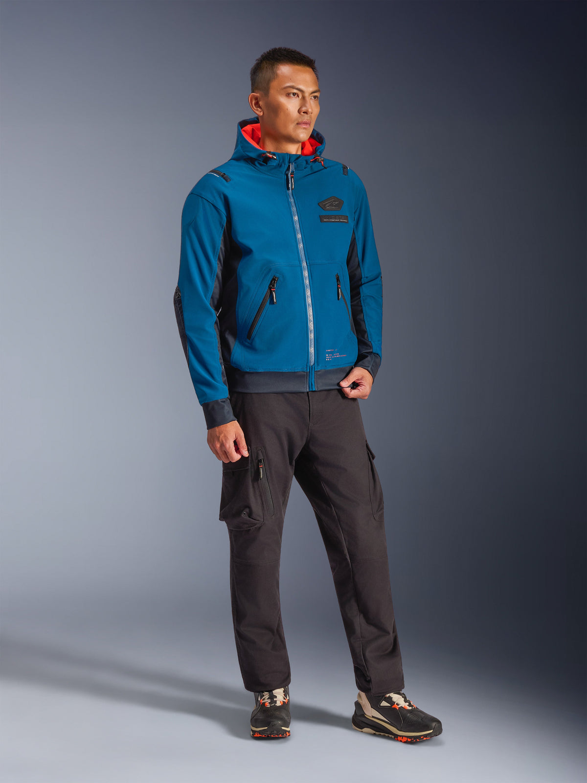 Männliches Model trägt die Alpinestars Moblast Waterproof Jacke in Dark Petrol und Dark Navy, mit Kapuzendesign und leuchtend orangefarbenem Innenfutter, kombiniert mit schwarzer Cargohose und Sneakern, moderne urbane Motorradbekleidung.