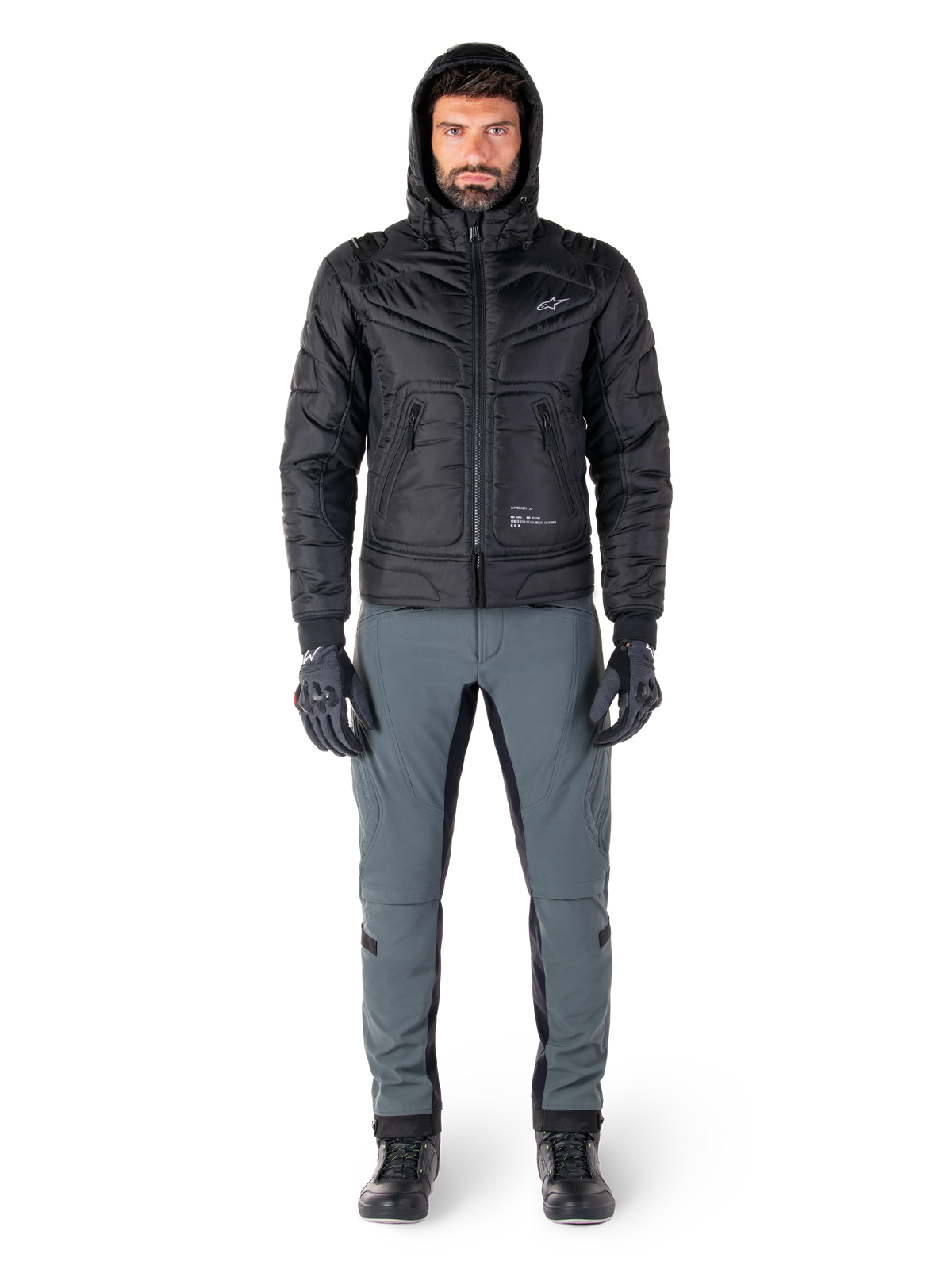 Alpinestars Mohobbs Wasserdichte Jacke in Schwarz, männliches Model in gesteppter wattierter Bomberjacke mit Kapuze, graue Motorradhose, schwarze Handschuhe und schwarze Stiefel, urbane zeitgenössische Motorradausrüstung.