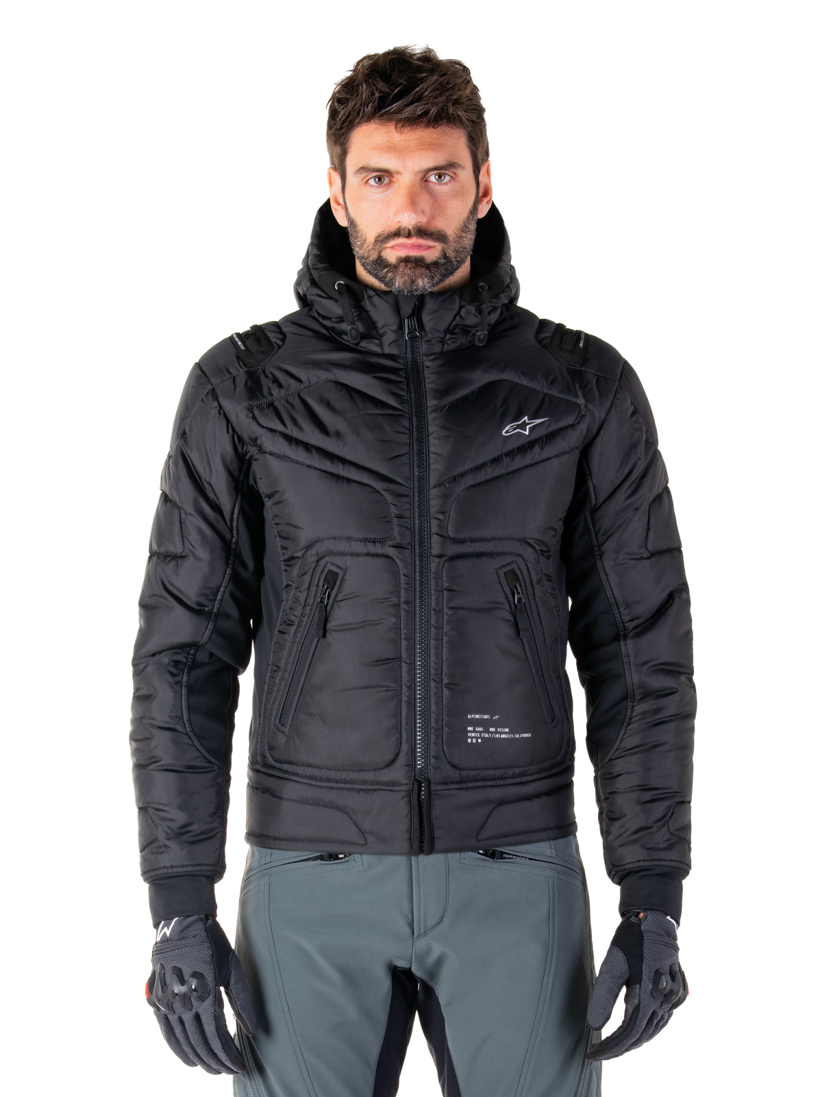 Alpinestars Mohobbs Waterproof Jacket, schwarze gepolsterte Bomber-Motorradjacke, gestepptes Polygewebe mit Frontreißverschluss und Reißverschlusstaschen, kleines weißes Logo auf der Brust, Modell trägt graue Hose und Handschuhe, urbanes Streetwear-Design für Stadtfahrten