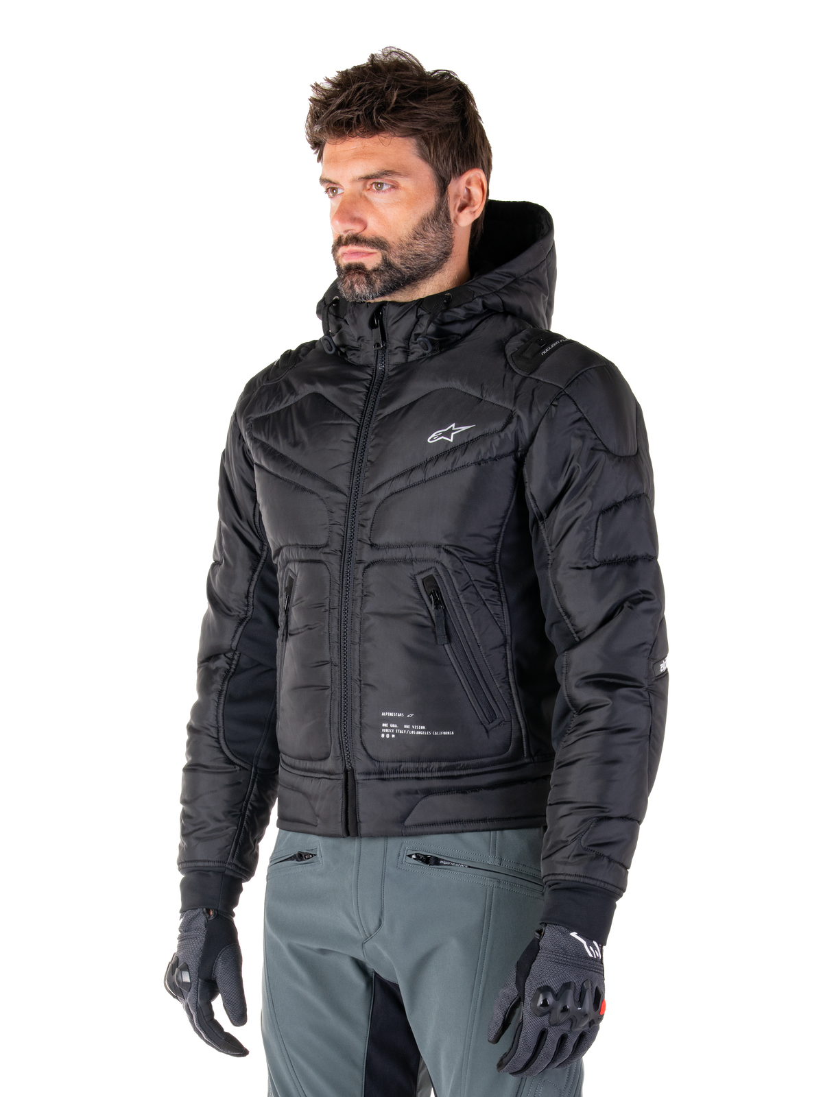 Alpinestars Mohobbs Wasserdichte Jacke, Schwarz, gestepptes wattiertes Bomberjacken-Design mit Kapuze, getragen von einem Mann mit grauer Motorradhose und schwarzen Handschuhen, urbane Streetwear-inspirierte Motorradbekleidung mit einem kleinen weißen Logo auf der Brust.