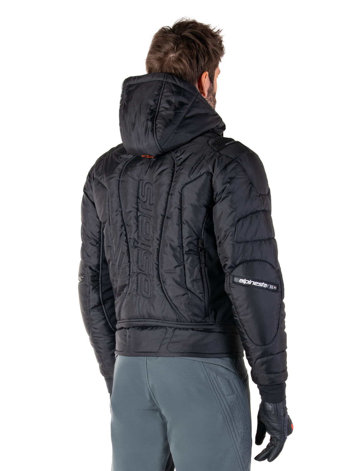 Alpinestars Mohobbs Wasserdichte Jacke, Schwarz, Rückansicht mit gesteppter wattierter Konstruktion, Kapuze, vertikaler Astars-Schriftzug auf dem Rücken und Alpinestars-Branding auf den Ärmeln, getragen mit grauer Hose und schwarzen Handschuhen.