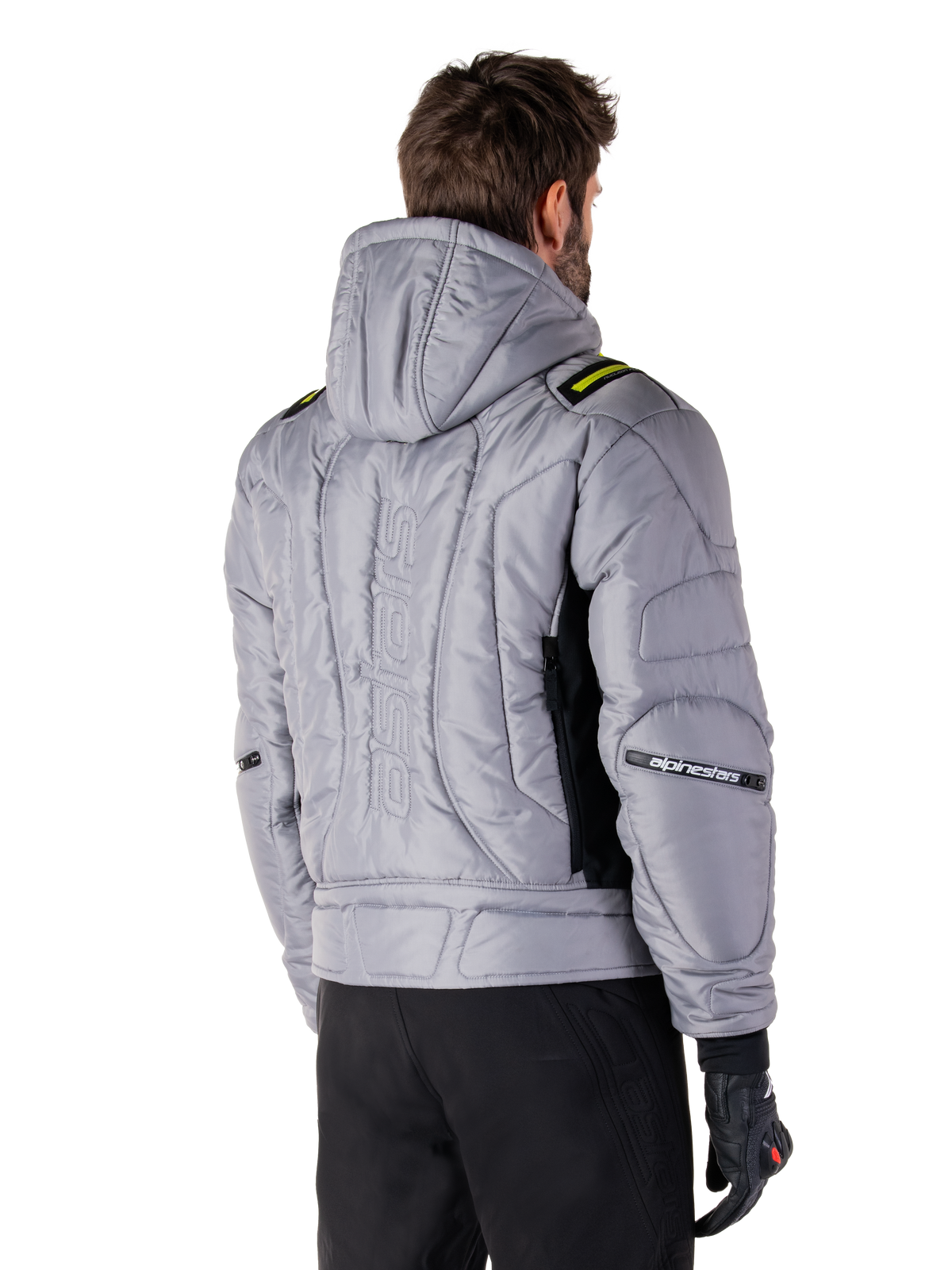 Alpinestars Mohobbs Waterproof Jas in Lunar Gray en Lime Punch, achteraanzicht op een model, gewatteerd bomberjack met verticaal astars-logo in reliëf op de rug, limegroene schouderaccenten, met capuchon, gedragen met zwarte motorbroek en handschoenen.