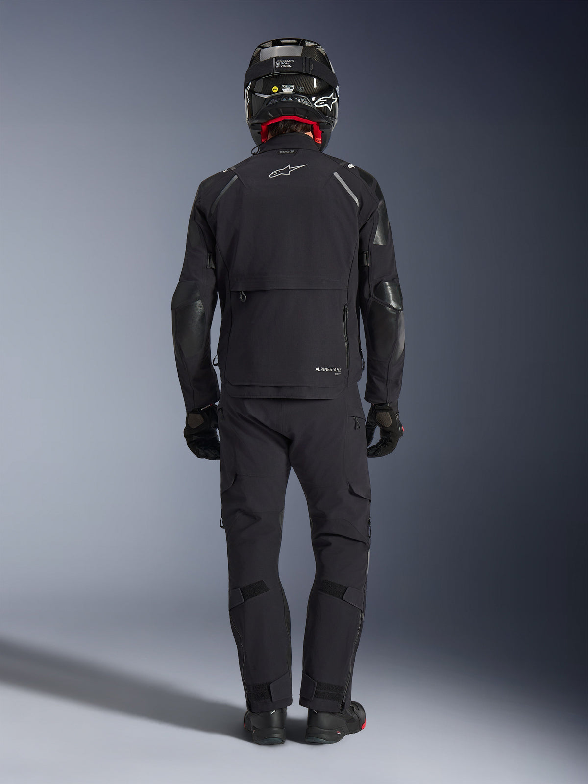 Alpinestars Andes Pro Drystar® XF jas in zwart en donkergrijs, achteraanzicht van een rijder in volledige adventure touring-uitrusting met bijpassende broek en carbon helm, voorzien van een groot opbergvak aan de achterzijde en reflecterende Alpinestars-branding op de rug.