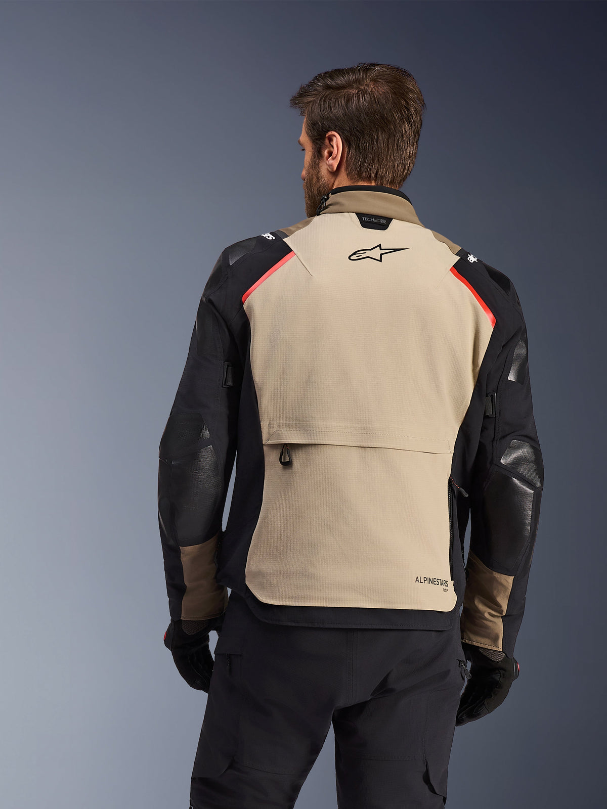Alpinestars Andes Pro Drystar® XF Jas, achteraanzicht, Walnut en Aluminum, voorzien van een groot beige rugpaneel met zwart Alpinestars logo en rode schouderaccenten, grote achterzak, gedragen met zwarte broek en handschoenen, adventure touring motoruitrusting
