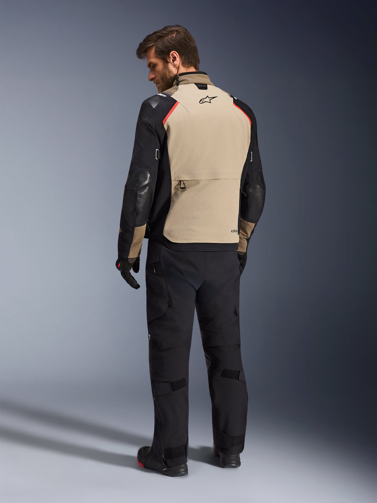 Alpinestars Andes Pro Drystar® XF Jacket, motorjas, walnoot en zwart met rode accenten, achteraanzicht op model, adventure touring ontwerp met grote opbergzak aan de achterzijde, gedragen met zwarte motorbroek en handschoenen.