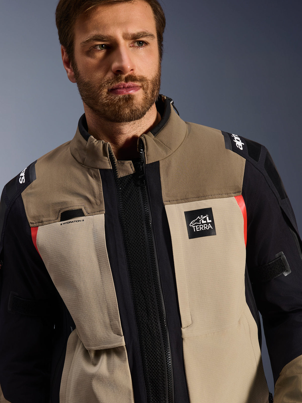 Alpinestars Andes Pro Drystar® XF jas in Walnut Aluminum, gedragen door een mannelijk model, adventure motorjas met een gelaagd textielontwerp in bruin, lichtgrijs en zwart met rode accenten, Alpinestars 'TERRA' logo op de borst en zichtbare technische details.