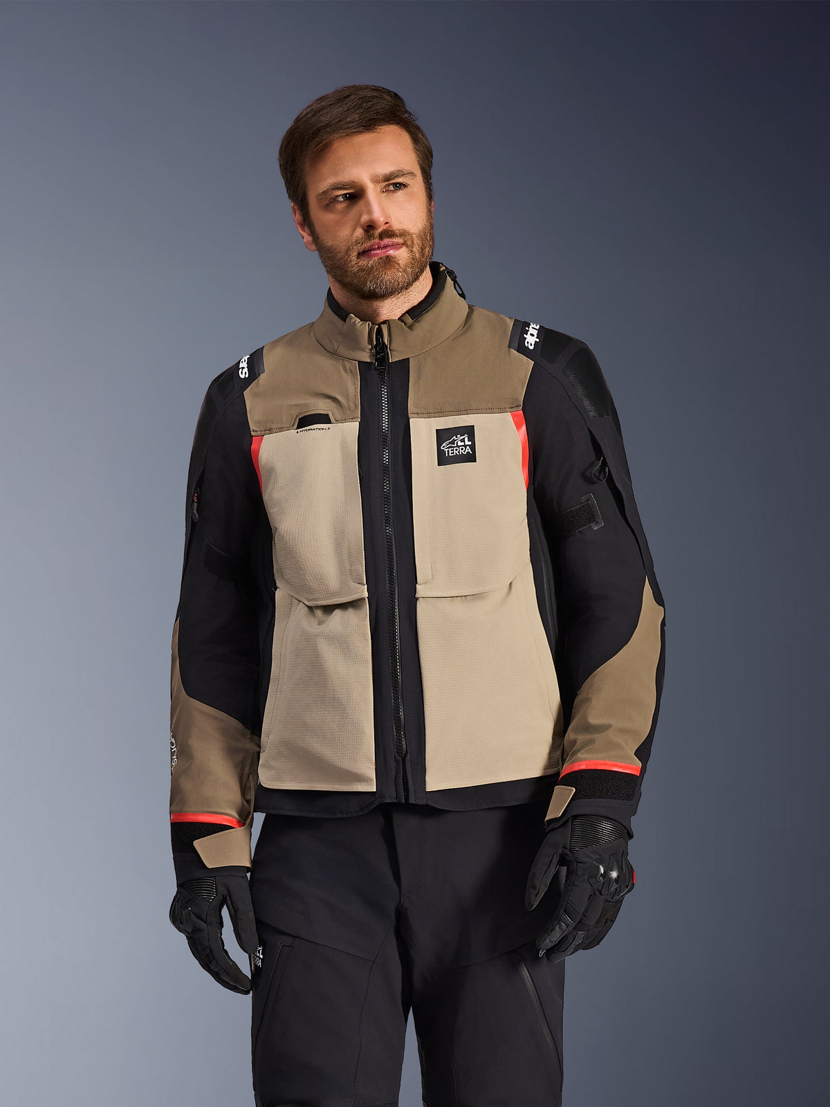 Alpinestars Andes Pro Drystar® XF Jas, Walnut Aluminum, adventure motorjas met beige en zwarte panelen en rode accenten, gedragen met een zwarte motorbroek en handschoenen, technische waterdichte touringuitrusting