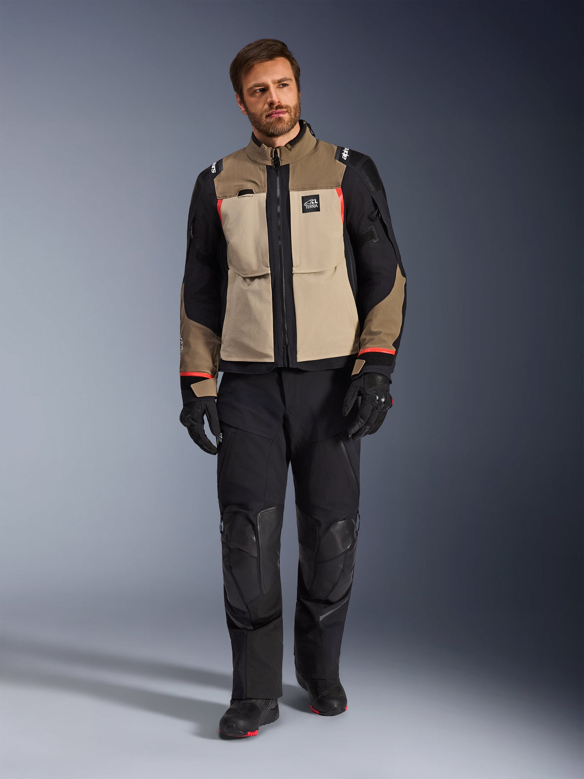 Mannelijk model draagt Alpinestars Andes Pro Drystar® XF Jacket in Walnut Aluminum, zandkleurige en zwarte adventure touring jas met rode accenten, zwarte motorbroek, handschoenen en laarzen, studio-opname van het volledige lichaam, beschermende motorkleding