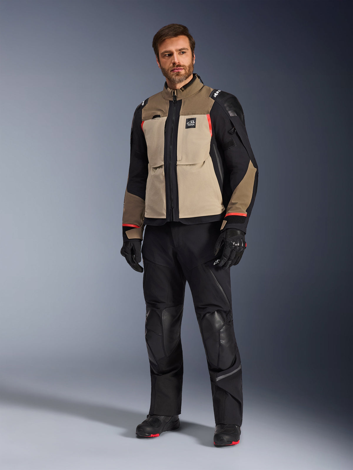 Mannelijk model in de Alpinestars Andes Pro Drystar® XF Jacket in Walnut Aluminum, gecombineerd met zwarte motorbroek, handschoenen en laarzen, met een tan en zwart multi-panel ontwerp met rode accenten voor adventure touring.