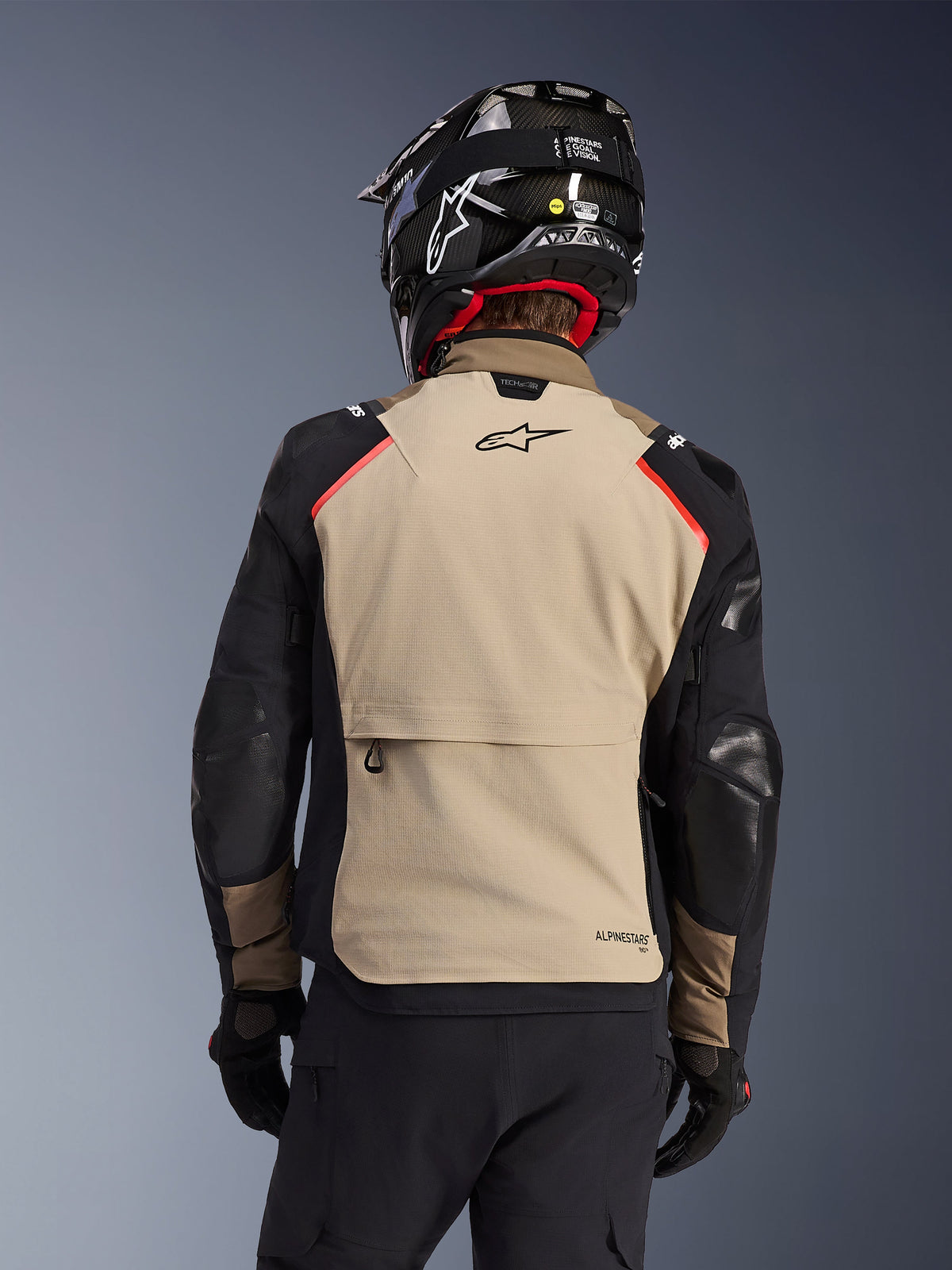Alpinestars Andes Pro Drystar® XF Jas in Walnut Aluminum, achteraanzicht, beige achterpaneel met grote cargozak, zwarte mouwen met versterkte panelen, Alpinestars-logo op de bovenrug, gecombineerd met een zwarte helm en handschoenen voor adventure touring.