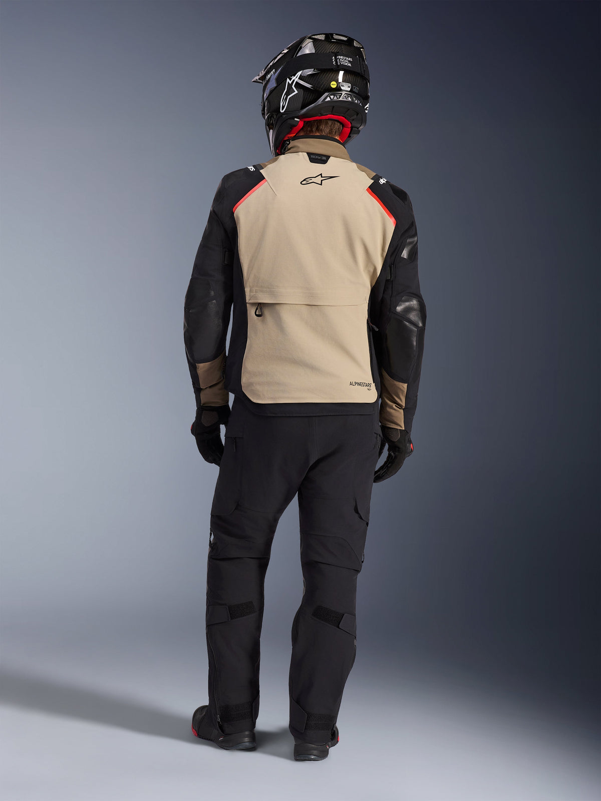 Rijder van achteren in de Alpinestars Andes Pro Drystar® XF Jas in Walnut Aluminum, beige rugpand met zwarte mouwen en rode accenten, gecombineerd met een zwarte technische adventure-broek en een motorhelm, hoogwaardige all-weather touringuitrusting.