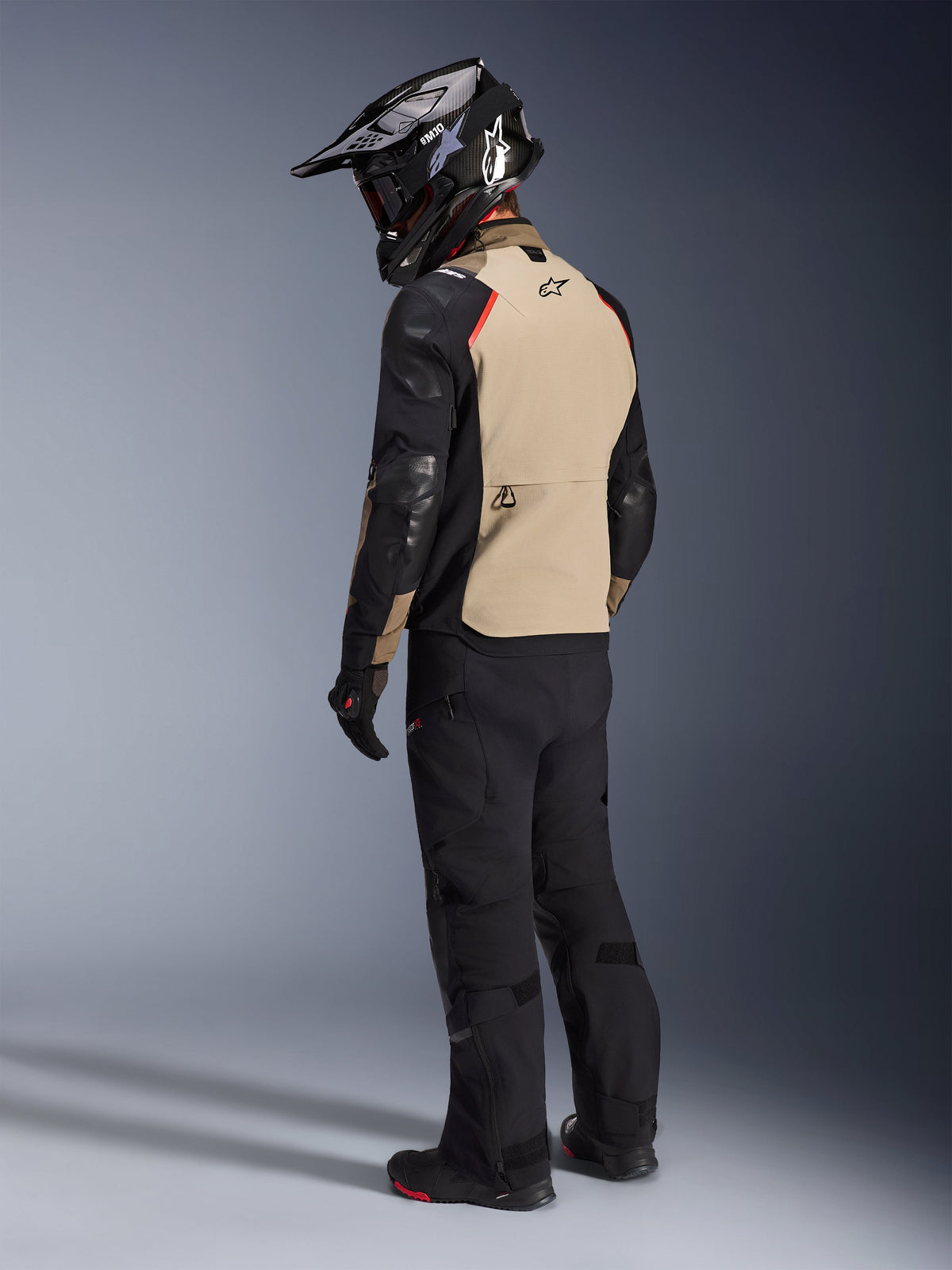 Alpinestars Andes Pro Drystar® XF Jas in Walnut Aluminum, motorrijder van achteren gezien, beige achterpand met zwarte mouwen en rode accenten, zwarte adventure-helm, zwarte motorbroek en zwarte laarzen, professionele adventure touring-uitrusting