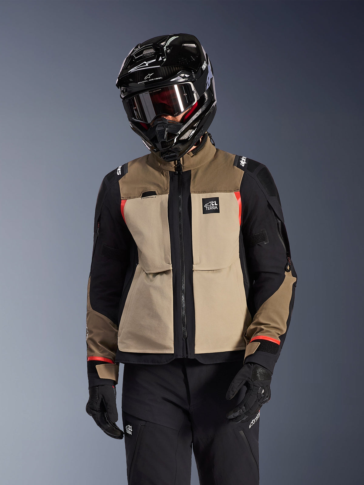 Alpinestars Andes Pro Drystar® XF jas, adventure touring motorjas, Walnut Aluminum beige en bruin met zwarte panelen en rode accenten, gedragen met een zwarte Alpinestars helm en motorbril, technische outdoor motorkleding