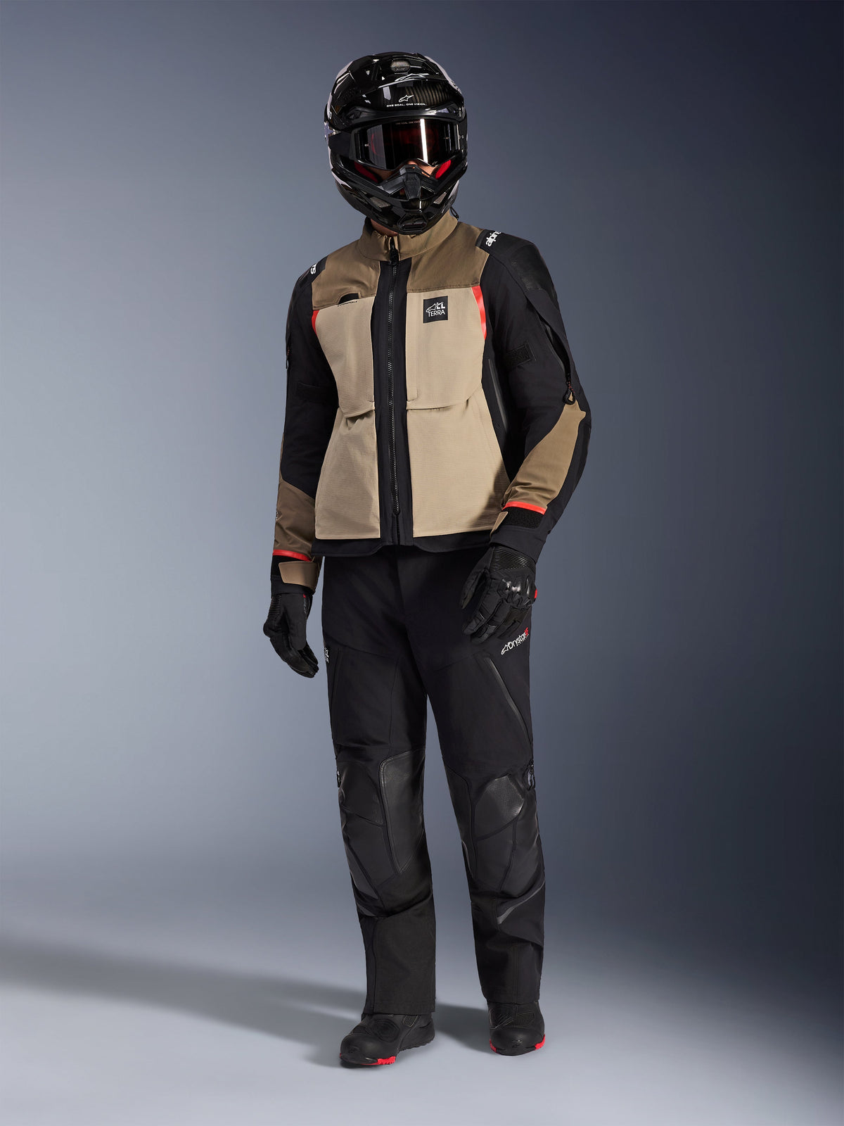 Alpinestars Andes Pro Drystar® XF jas in Walnut Aluminum, adventure motorjas gedragen met bijpassende zwarte broek, een zwarte adventure-helm met bril en zwarte laarzen, voorzien van beige en zwarte panelen met rode accenten.