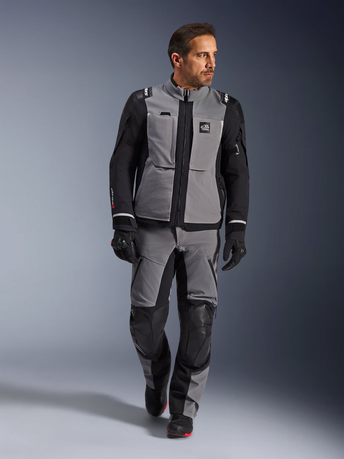Modèle masculin portant la veste Alpinestars Andes Pro Drystar® XF en gris foncé et noir, tenue de moto aventure avec pantalon et gants assortis, dotée d'une coque technique en textile avec marquage « All Terra », équipement complet pour le touring d'aventure longue distance.