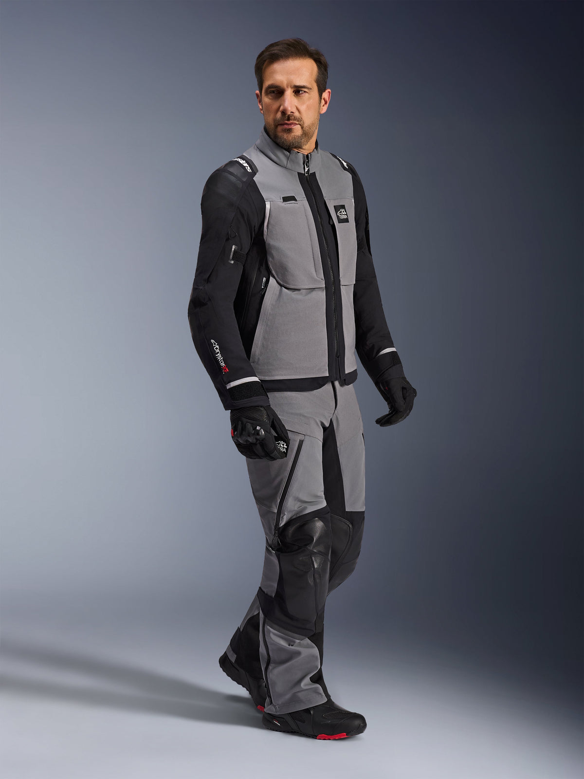 Veste Alpinestars Andes Pro Drystar® XF, veste de moto d'aventure, gris foncé et noir, portée par un mannequin avec un pantalon en textile assorti et des gants noirs, dotée d'une conception multi-poches, d'une coque laminée technique à 3 couches et d'une coupe articulée pour les longs trajets d'aventure.