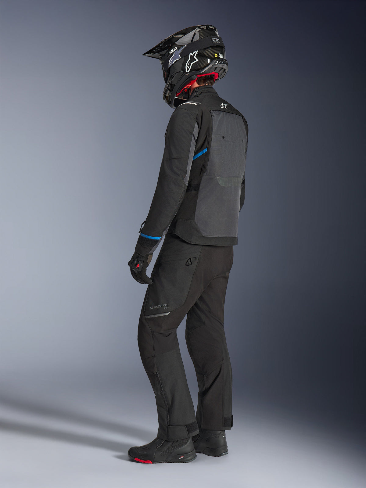 Motorcycle rider wearing the Alpinestars Cusco Drystar® Jacket in Black and Deep Gray with blue accents, paired with black riding pants, a carbon fiber SM10 helmet, and black gloves, back view showing the large ventilation panel and adventure touring design.