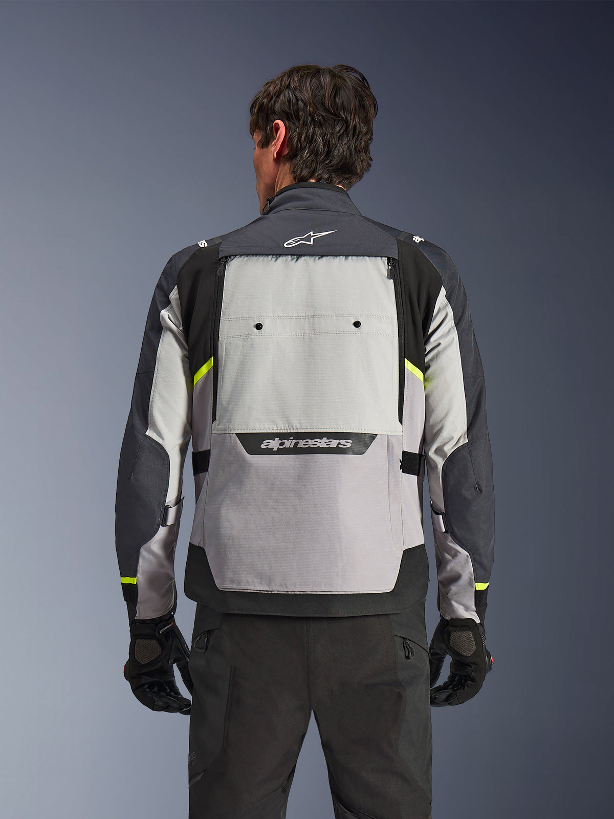 Rear view of a rider wearing the Alpinestars Cusco Drystar® Jacket in Ice Gray, Mid Gray, and Deep Gray, featuring neon yellow accents, a large back ventilation panel, and bold Alpinestars branding, paired with black riding pants and gloves.