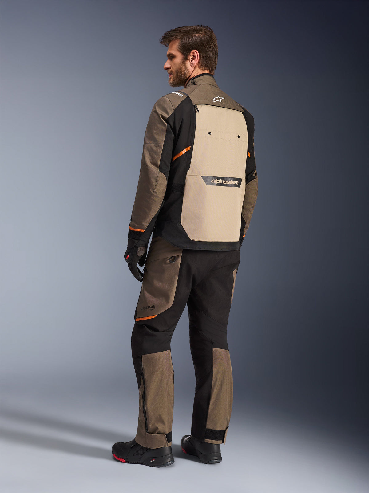 Alpinestars Cusco Drystar® Jas in Walnut Aluminum Black, achteraanzicht met beige rugpaneel en oranje accenten, gedragen met bijpassende motorbroek, zwarte handschoenen en laarzen, professionele adventure touring uitrusting