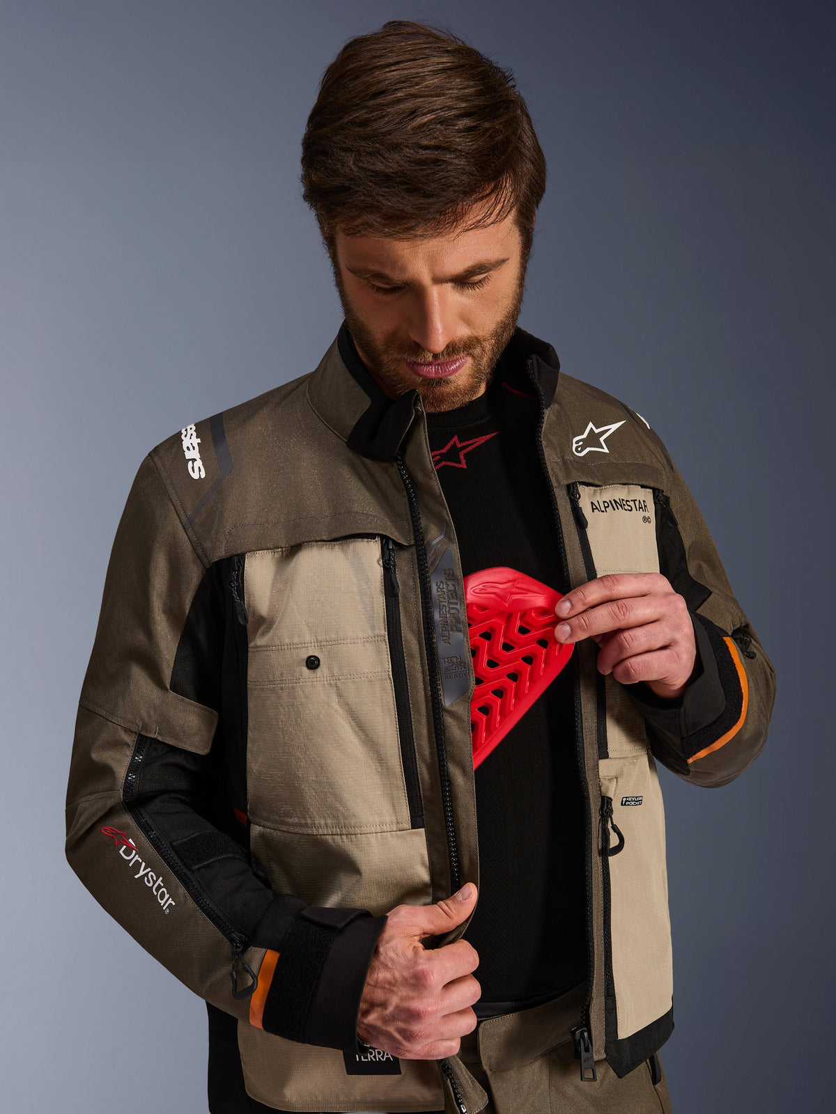 Alpinestars Cusco Drystar® Jacket, Walnut Aluminum Black, gedragen door een man die het borstbeschermingsvak toont met een rood protectie-inzetstuk, textiele motorjas met ventilatieopeningen met ritssluiting en branding voor adventure touring.