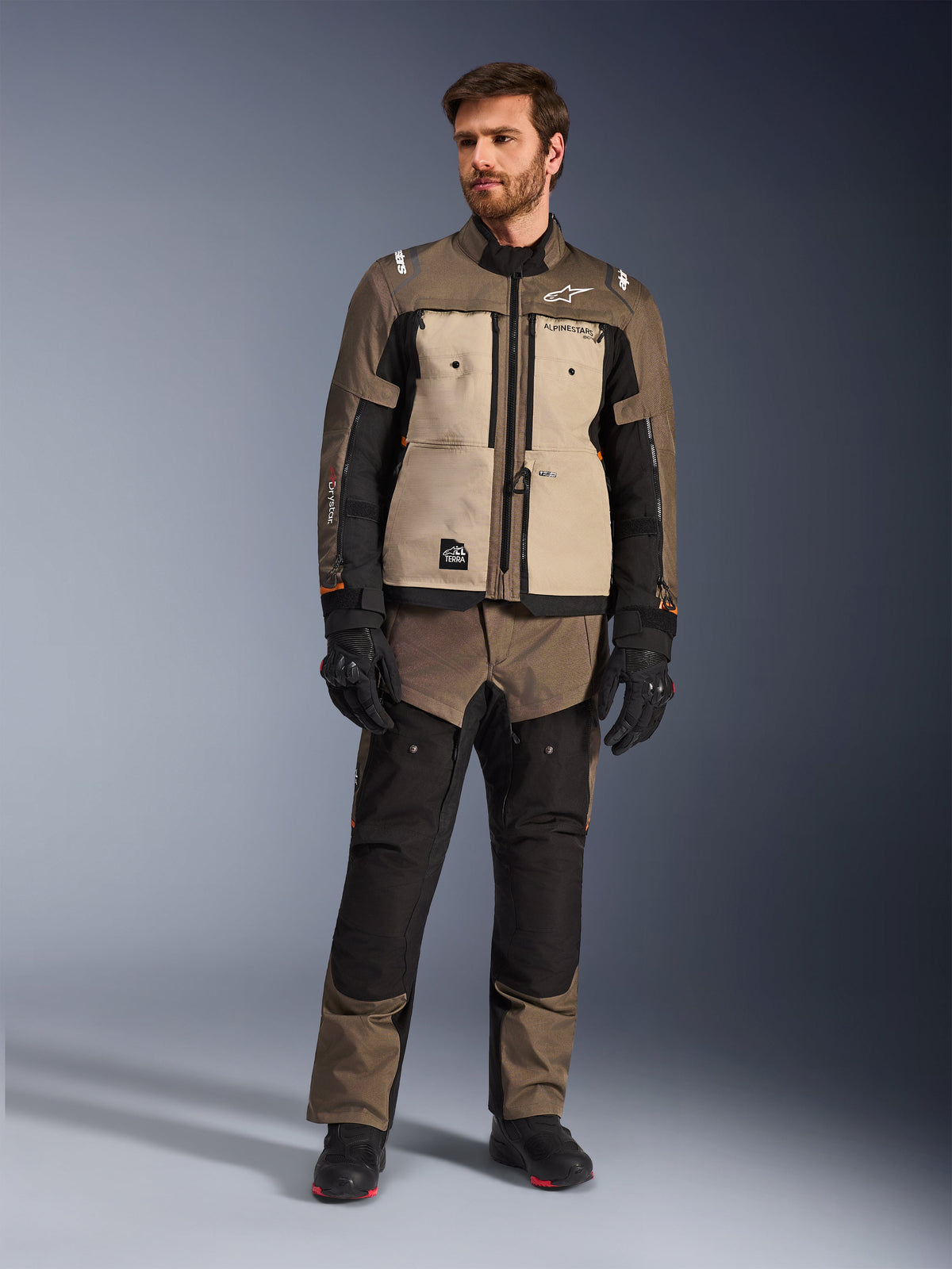 Alpinestars Cusco Drystar® Jacket in Walnut Aluminum Black, gedragen door een mannelijke motorrijder met bijpassende adventure touring broek, zwarte handschoenen en laarzen, voorzien van zandkleurige borstpanelen, zwarte mouwen met subtiele oranje accenten en functionele ventilatieritsen voor motorrijden op de weg.