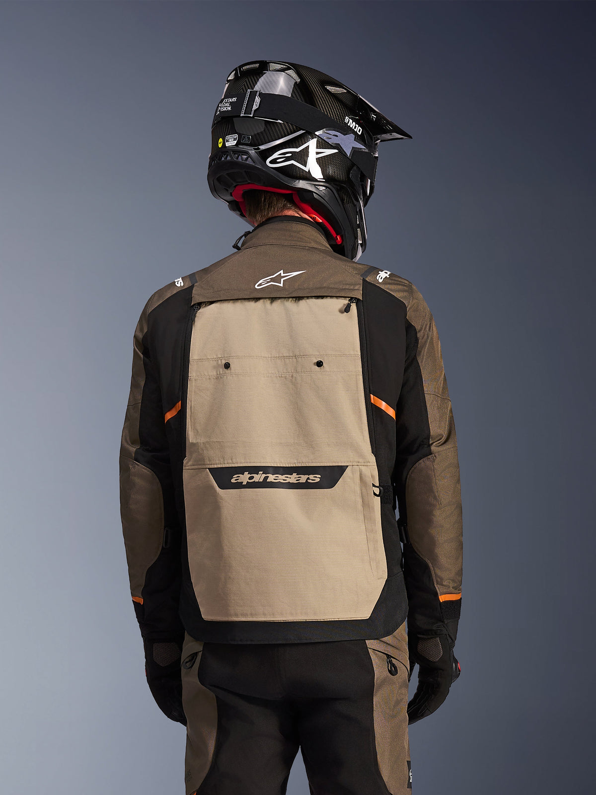 Achteraanzicht van de Alpinestars Cusco Drystar® jas in Walnut Aluminum Black, adventure touring motorjas met een groot beige rugpaneel met Alpinestars-branding, zwarte mouwen met oranje accenten, getoond met een zwarte carbon-stijl helm.