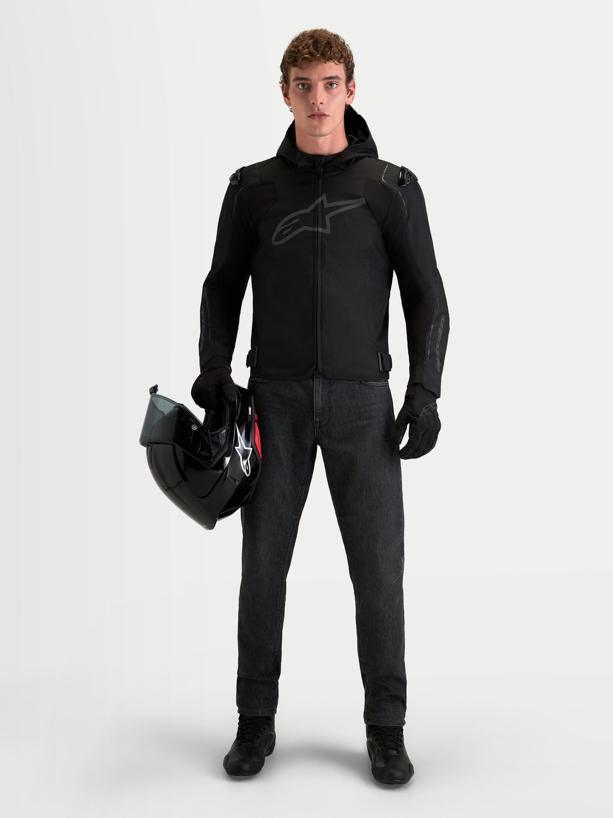 Alpinestars Zaca V 2 Waterproof Veste, veste de moto noire avec capuche et protections d'épaules, portée par un homme tenant un casque