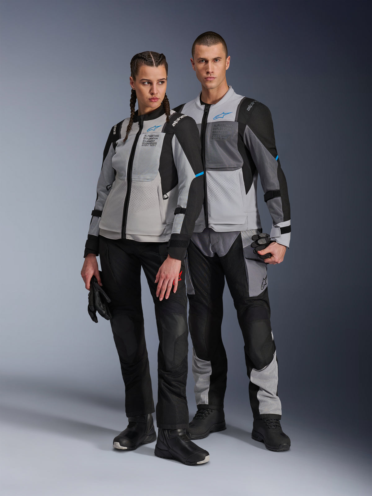 Man en vrouw in Alpinestars Stella ST-2 Air jassen en bijpassende technische broeken, Ice Gray en zwarte kleurstelling met blauwe accenten, voorzien van mesh-ventilatie en 'ALPINESTARS EXPLORATION EQUIPMENT' branding, studiofoto van koppel