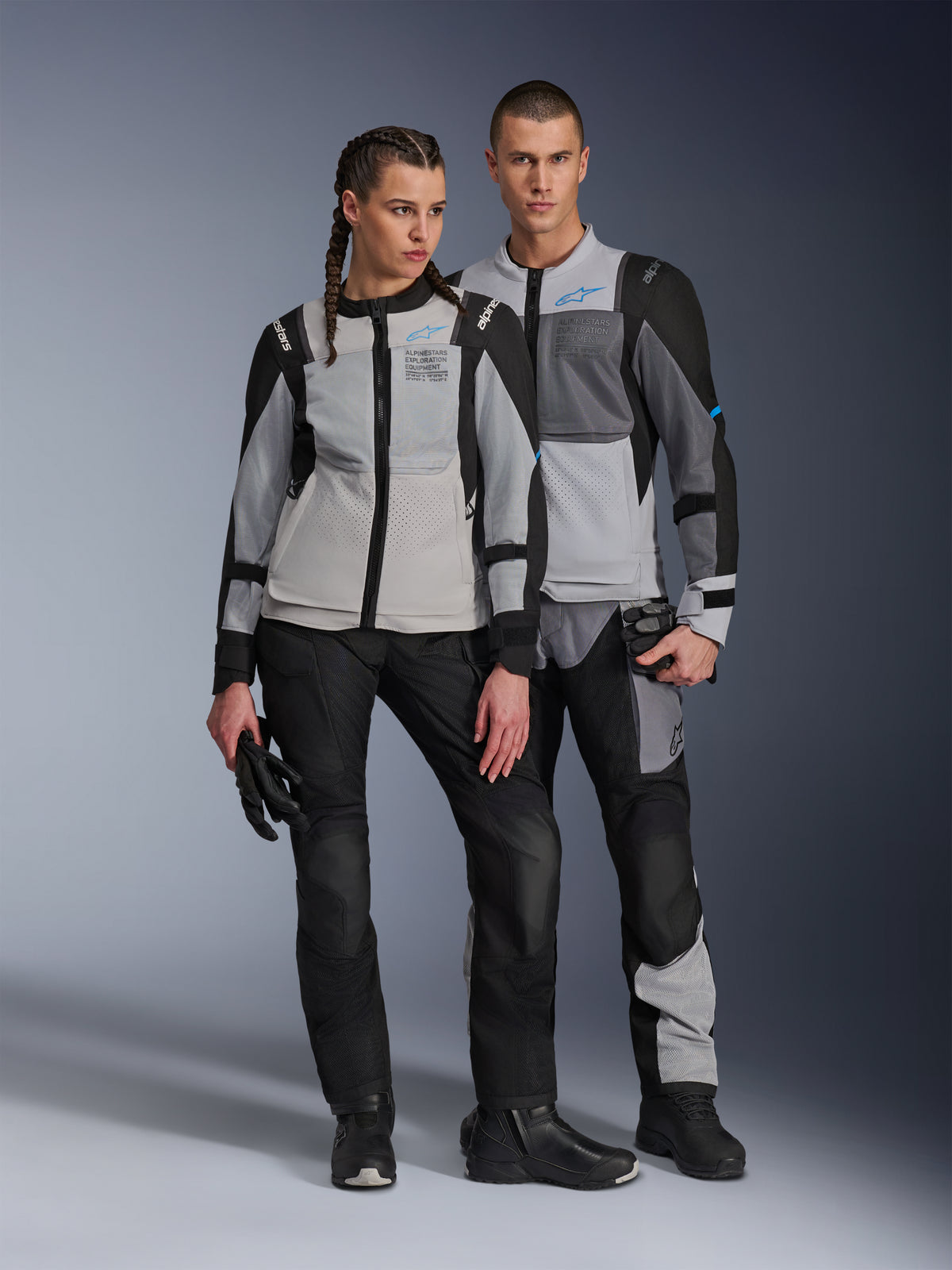 Männliches und weibliches Model in Alpinestars ST-2 Air Jacken in Ice Gray, Dark Gray und Schwarz, technische Sommer-Touring-Motorradausrüstung mit großen Mesh-Einsätzen und Exploration Equipment-Branding, kombiniert mit schwarzen Motorradhosen, Handschuhen und Stiefeln.