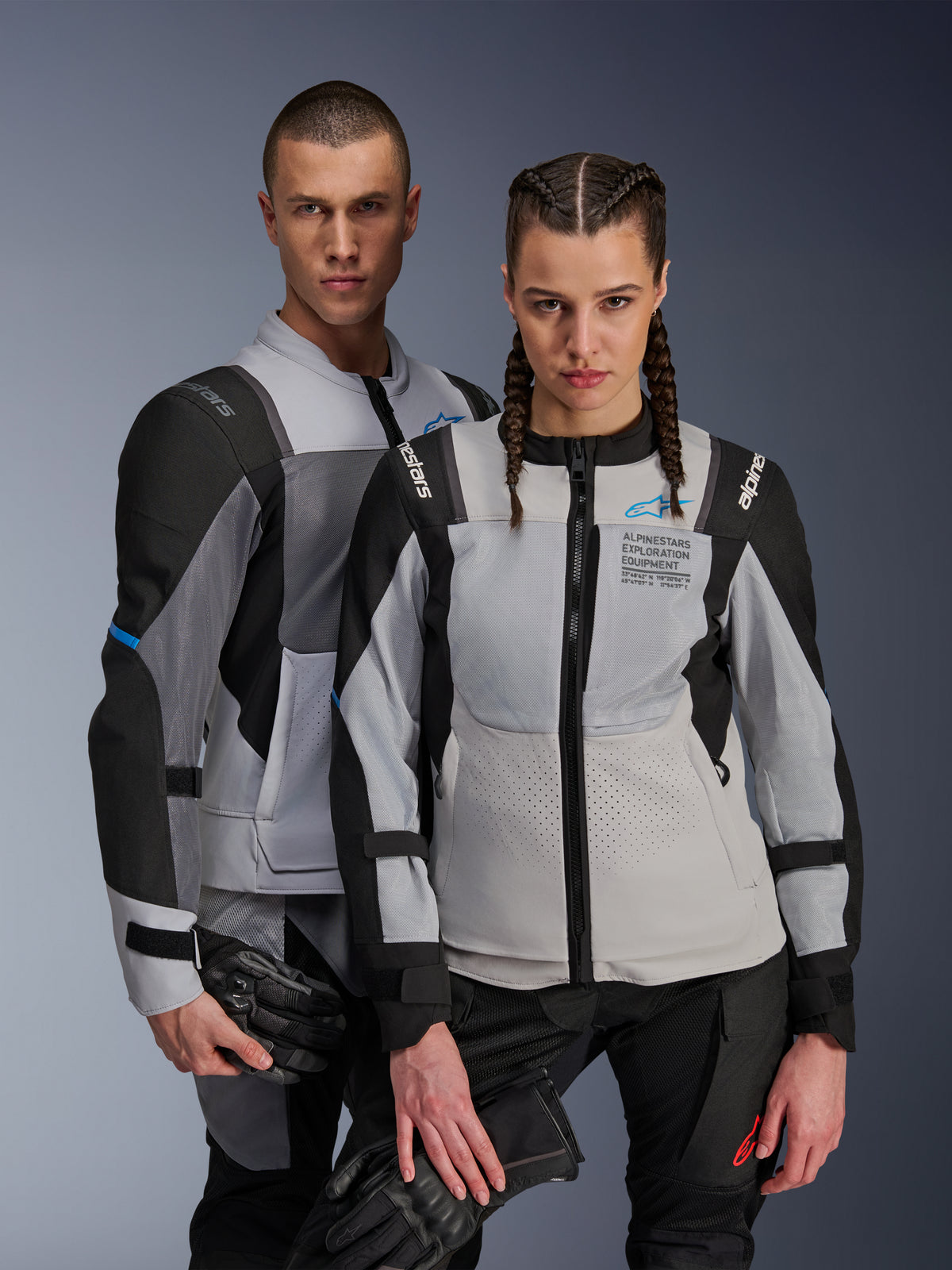 Alpinestars ST-2 Air en Stella ST-2 Air jassen, Ice Gray en zwart, textiele motorjassen met meshpanelen en laserperforaties, voorzien van 'Alpinestars Exploration Equipment' branding, gedragen door een mannelijk en vrouwelijk model die motorhandschoenen vasthouden.