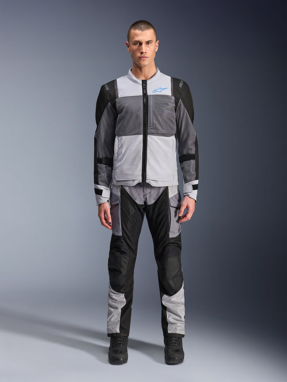 Alpinestars ST-2 Air Jacke, Motorrad-Tourenjacke, Eisgrau, Dunkelgrau und Schwarz, getragen von einem Modell mit passender Adventure-Hose, mit großen Mesh-Einsätzen, blauem Alpinestars-Logo auf der Brust und Alpinestars Exploration Equipment-Branding.