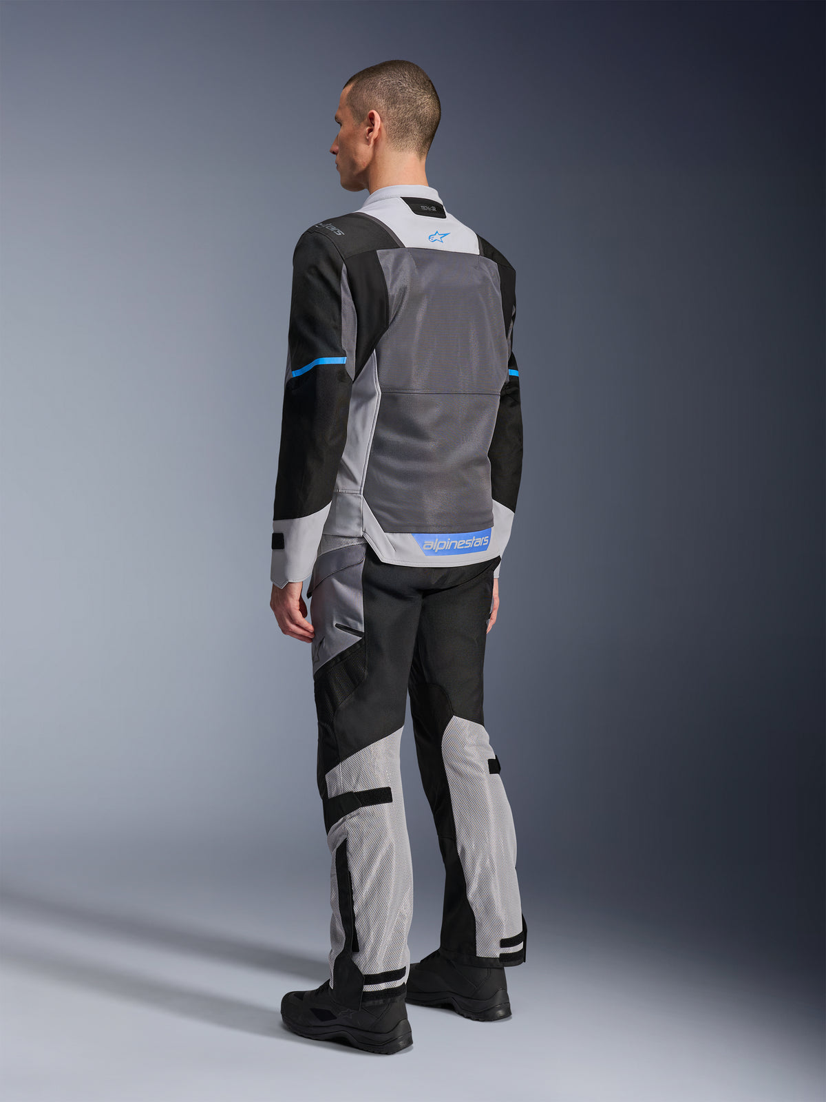 Männliches Model von hinten in der Alpinestars ST-2 Air Jacke in Ice Gray, Dark Gray und Schwarz, mit einem großen grauen Mesh-Rückeneinsatz, blauen Logo-Akzenten und passender Motorradhose für Sommertouren.