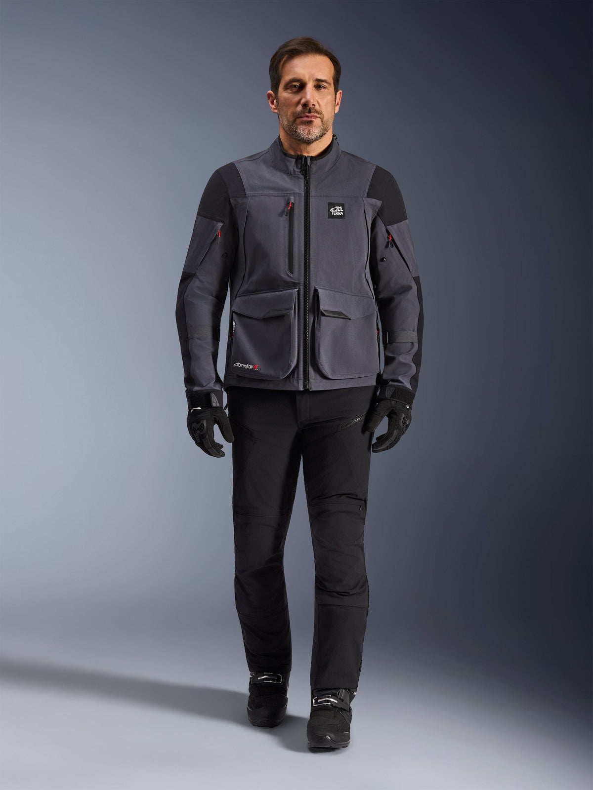 Maxdura Drystar®XF Laminated Armored Shell, motorjas, donkergrijs zwart, gedragen door een model met zwarte broek en handschoenen, voorzien van grote cargozakken, rode ritsaccenten en All Terra-branding, technische adventure rijuitrusting