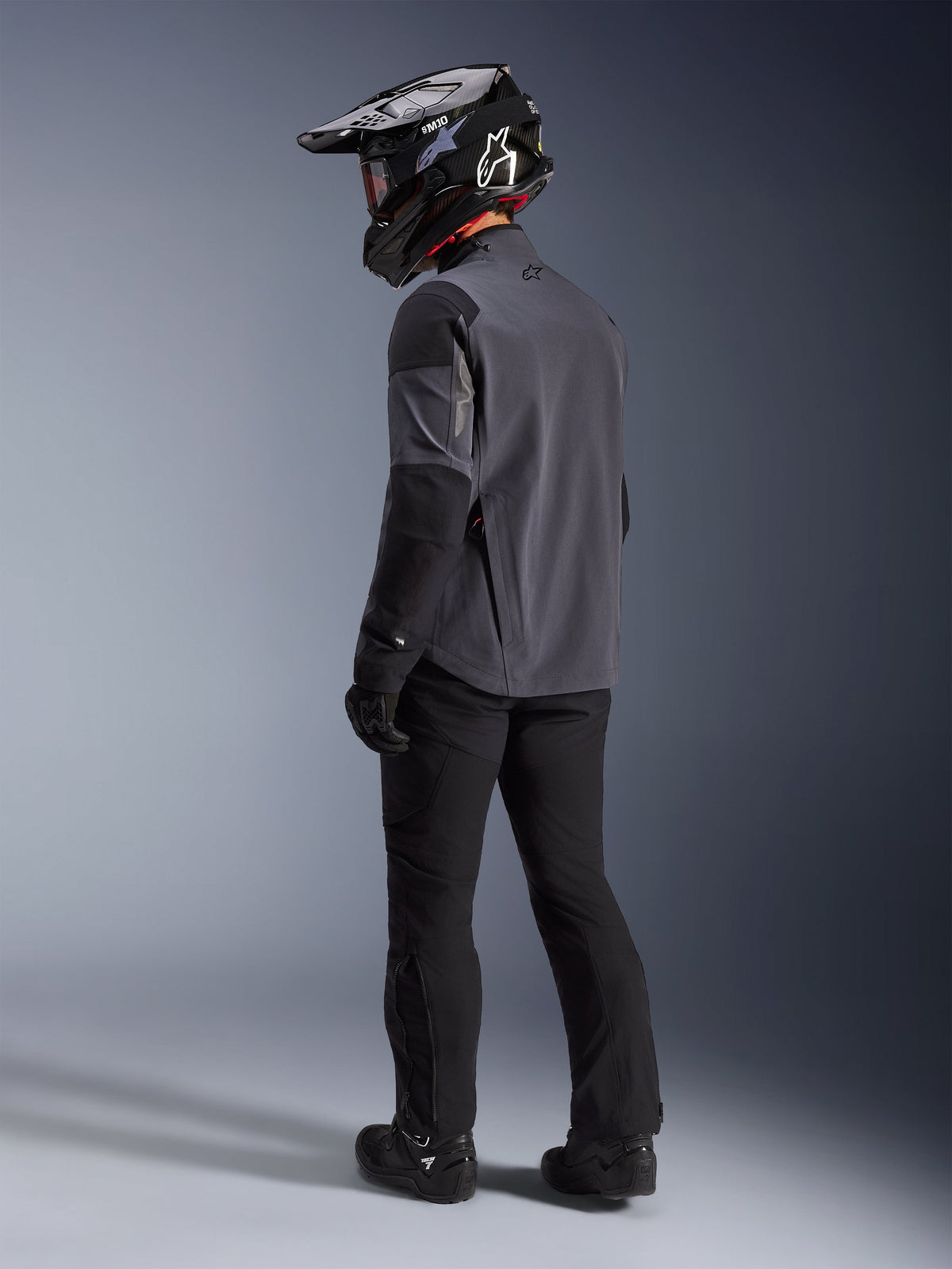 Adventure-rijder in de Maxdura Drystar®XF Laminated Armored Shell in Deep Gray Black, achteraanzicht. Complete uitrusting inclusief zwarte motorbroek, zwarte motorcrosshelm met Alpinestars-branding en zwarte off-road laarzen. Technische all-weather rijuitrusting voor dual-sport prestaties.