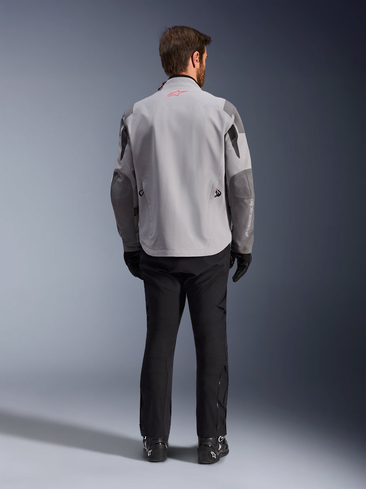 Alpinestars Maxdura Drystar®XF Laminated Armored Shell, motorjas in Mid Gray en Dark Gray, achteraanzicht, met een lichtgrijze body en donkergrijze panelen op de schouders en mouwen, klein rood Alpinestars sterlogo op de achterkant van de kraag en witte merknaam op de linker ondermouw, gecombineerd met een zwarte motorbroek en laarzen.