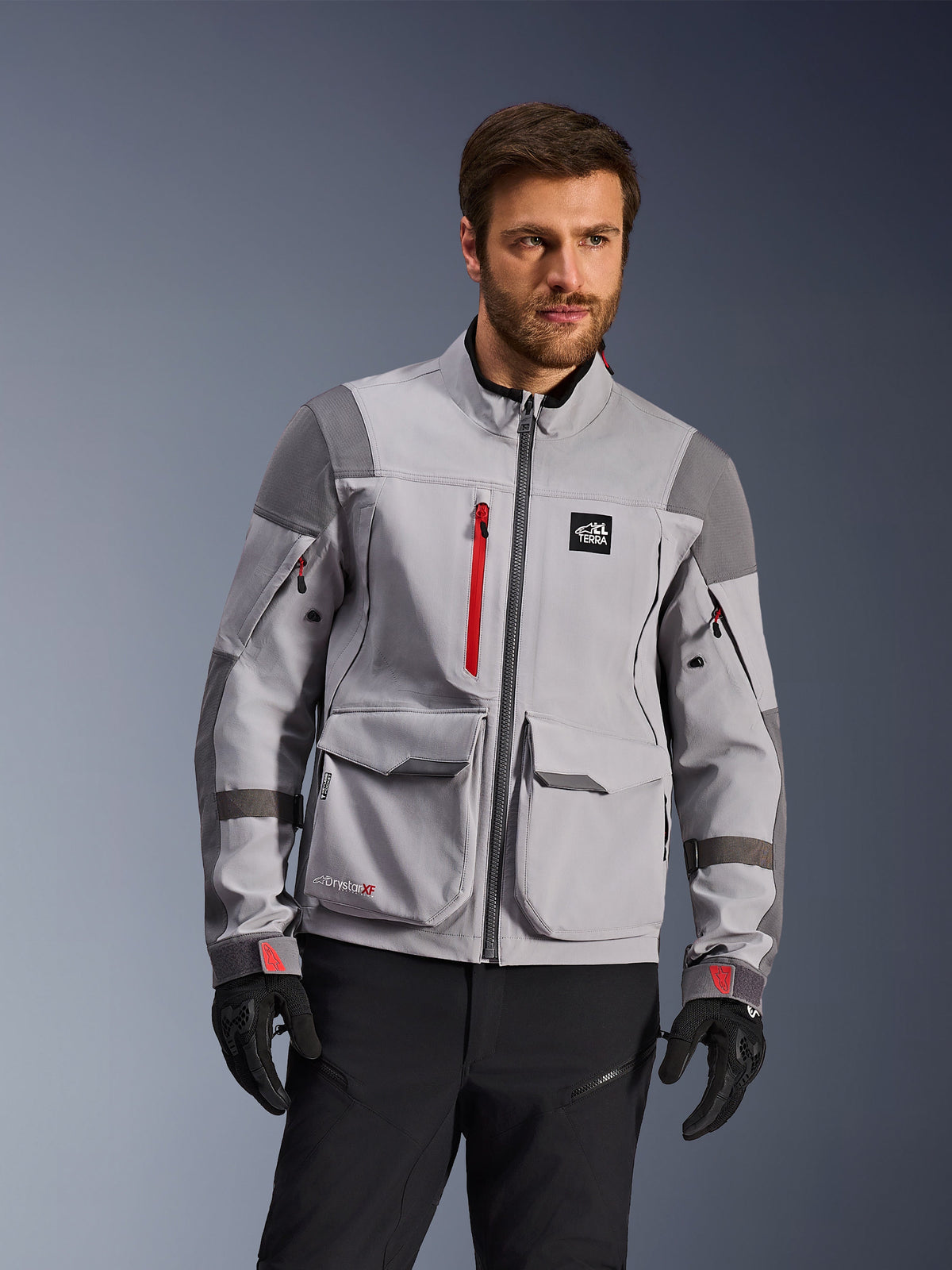 Alpinestars Maxdura Drystar®XF gelamineerde shell met protectie, adventure motorjas, middengrijs en donkergrijs met rode accenten, gedragen door een model met zwarte handschoenen en broek, voorzien van grote cargozakken en waterdichte ritsen, professionele motorkleding