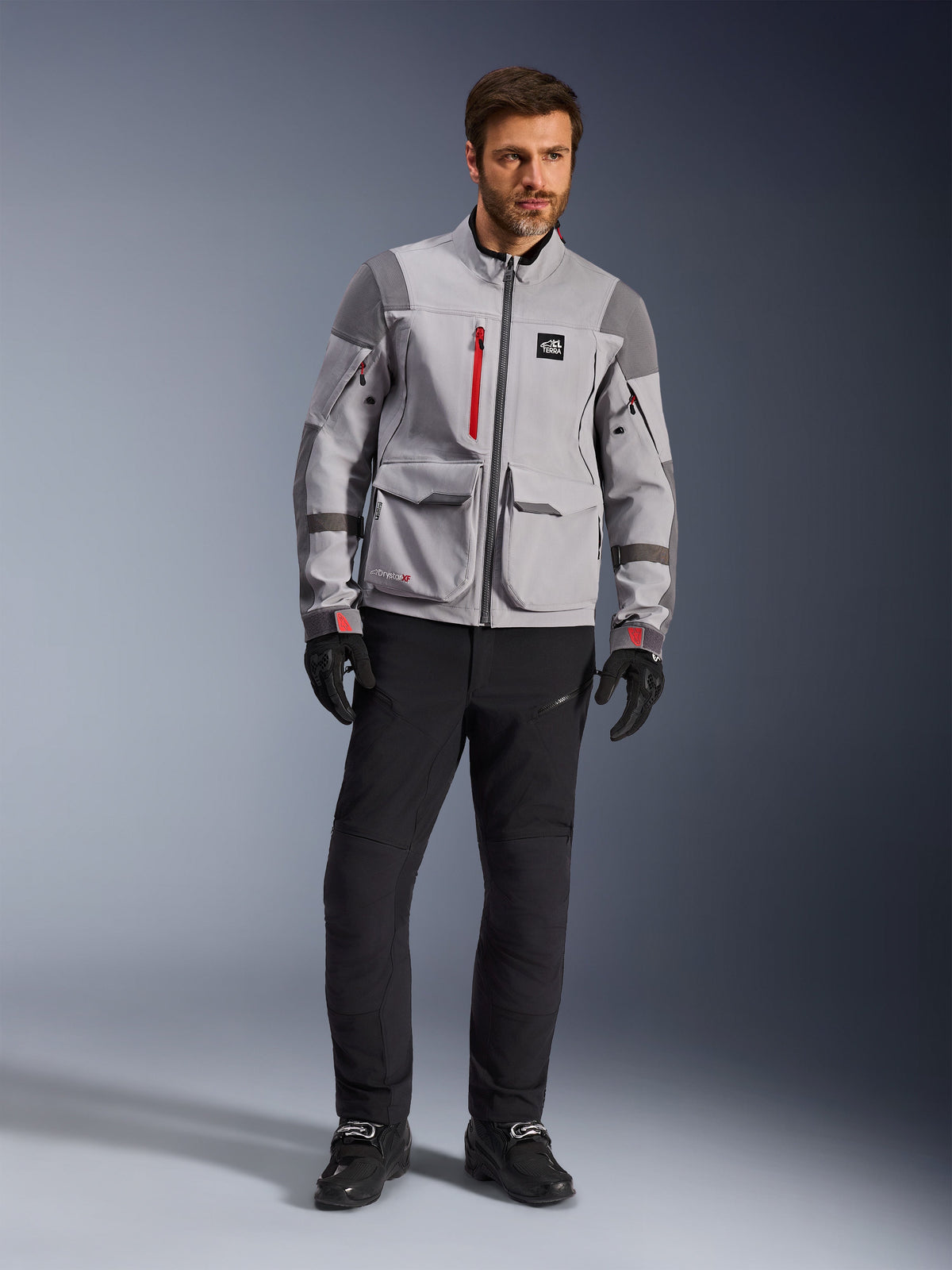 Alpinestars Maxdura Drystar®XF Gelamineerde Gepantserde Shell, adventure motorjas, middengrijs en donkergrijs met rode accenten, gedragen met zwarte motorbroek en handschoenen, vooraanzicht met nadruk op de technische constructie en grote cargozakken.