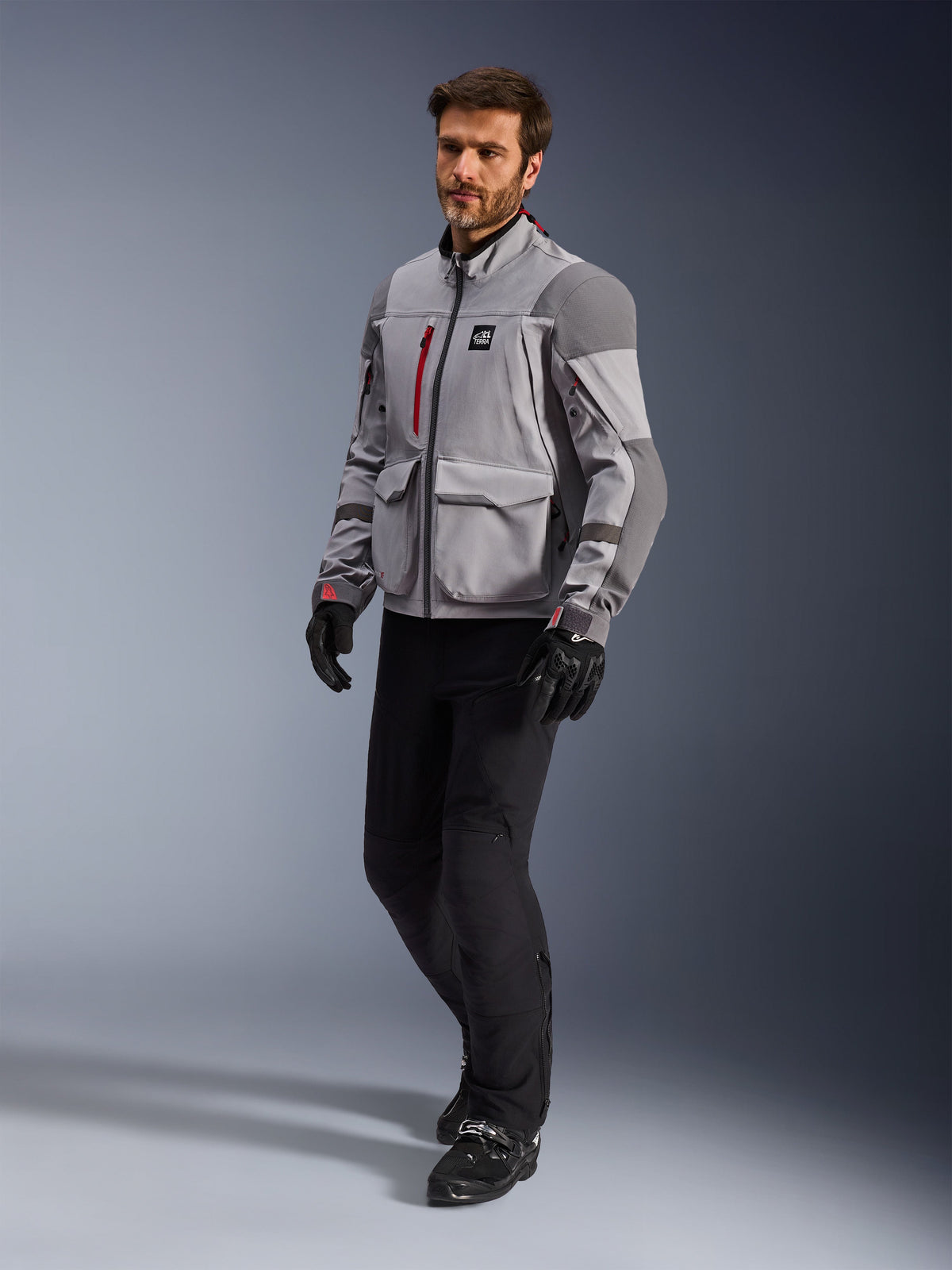 Alpinestars Maxdura Drystar®XF Gelamineerde Gepantserde Shell, adventure motorjas, middengrijs en donkergrijs, gedragen door een model met zwarte motorbroek en handschoenen, voorzien van rode ritsaccenten en cargozakken, professionele technische uitrusting voor alle weersomstandigheden voor adventure touring