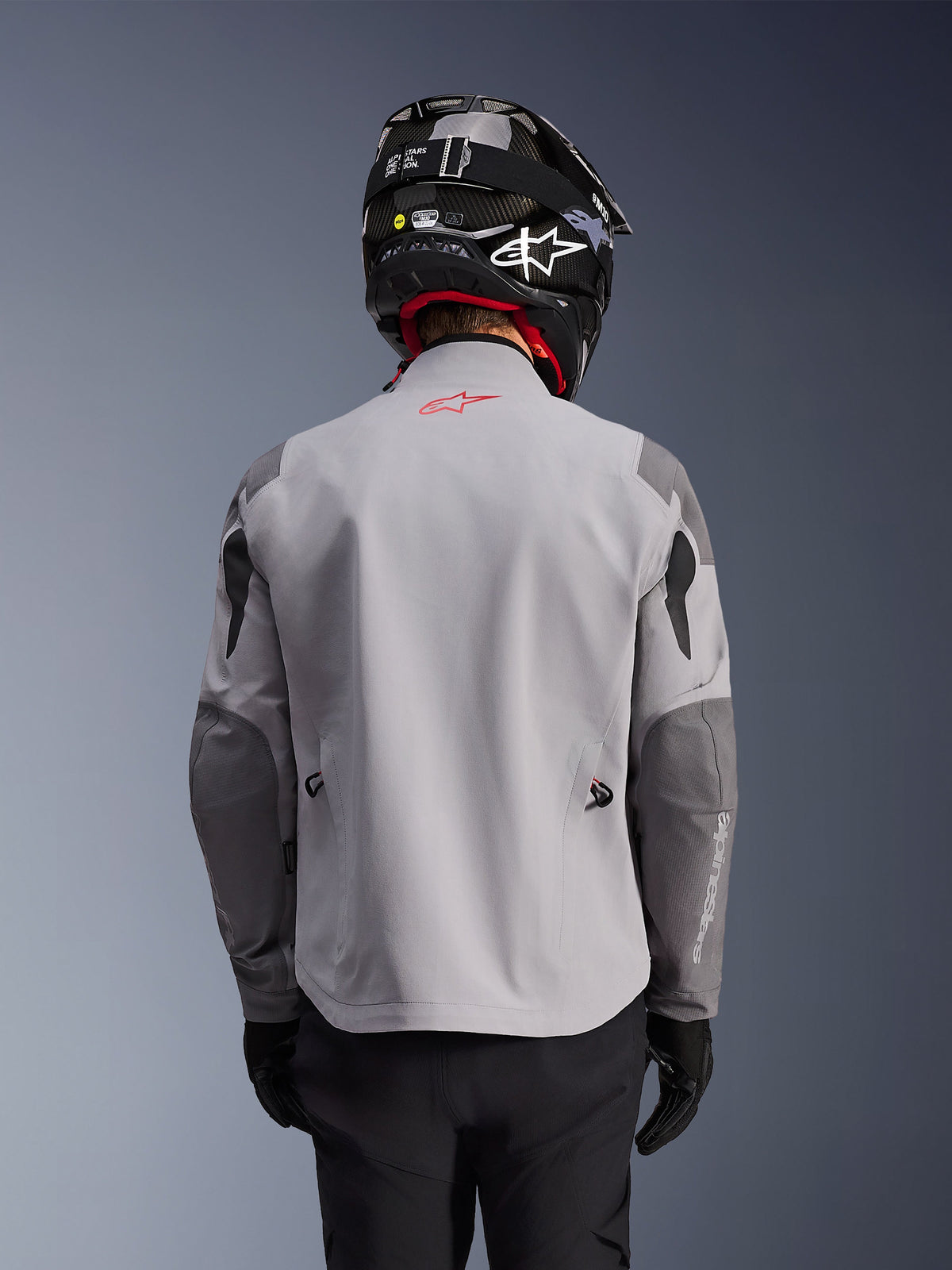 Alpinestars Maxdura Drystar®XF Gelamineerde Gepantserde Shell, adventure motorjas, middengrijs en donkergrijs, achteraanzicht met lichtgrijze body en donkergrijze versterkte panelen op schouders en ellebogen, rood Alpinestars-logo op de bovenrug, gedragen met een zwarte helm en zwarte handschoenen.