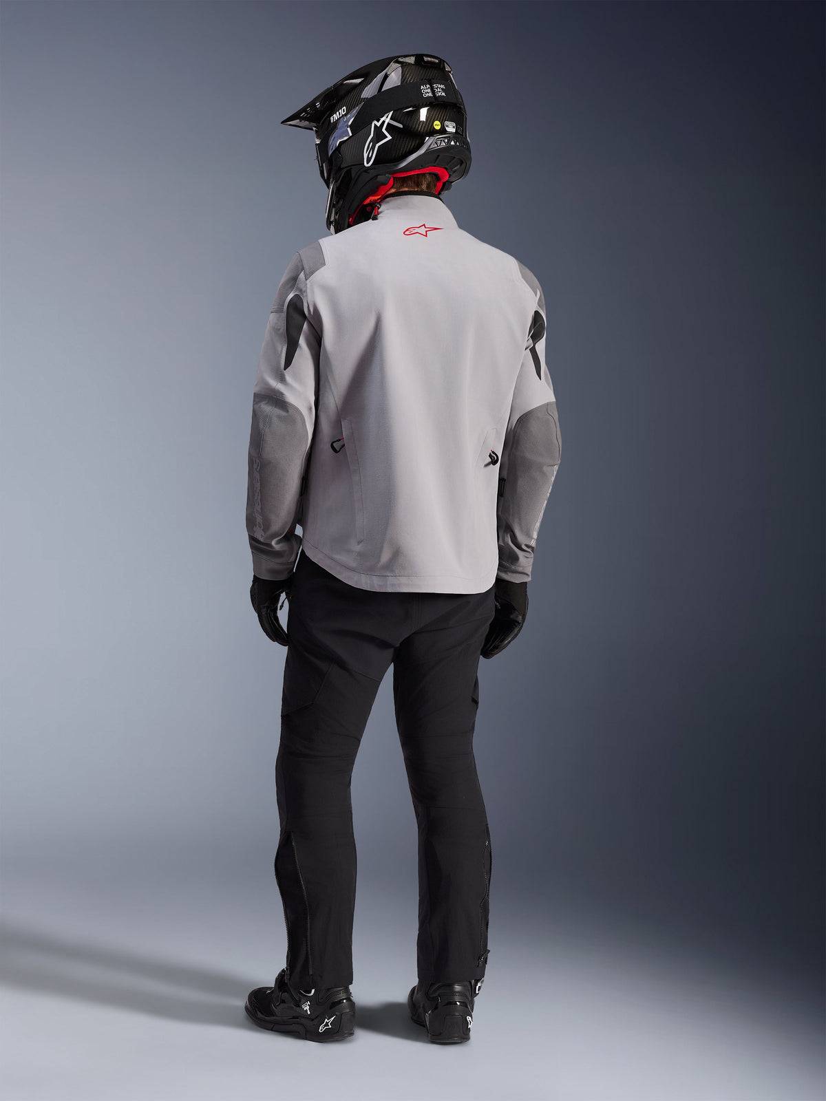 Achteraanzicht van een motorrijder die de Alpinestars Maxdura Drystar®XF Laminated Armored Shell-jas draagt in Mid Gray en Dark Gray, voorzien van een rood sterlogo op de kraag, gecombineerd met een zwarte adventure-broek, een zwarte helm met carbonpatroon en zwarte laarzen.