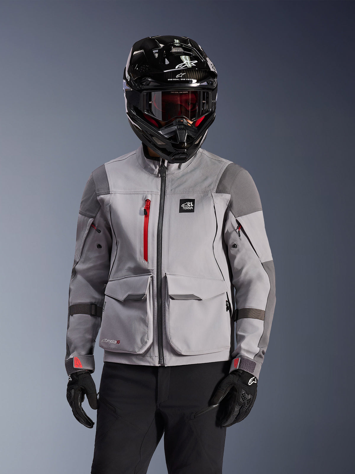 Alpinestars Maxdura Drystar®XF Laminated Armored Shell, adventure motorjas in Mid Gray en Dark Gray, gedragen met een zwarte helm met carbon-afwerking en een rood getinte bril, voorzien van cargozakken aan de voorzijde en een rode rits op de borst, technisch ontwerp voor adventure touring.