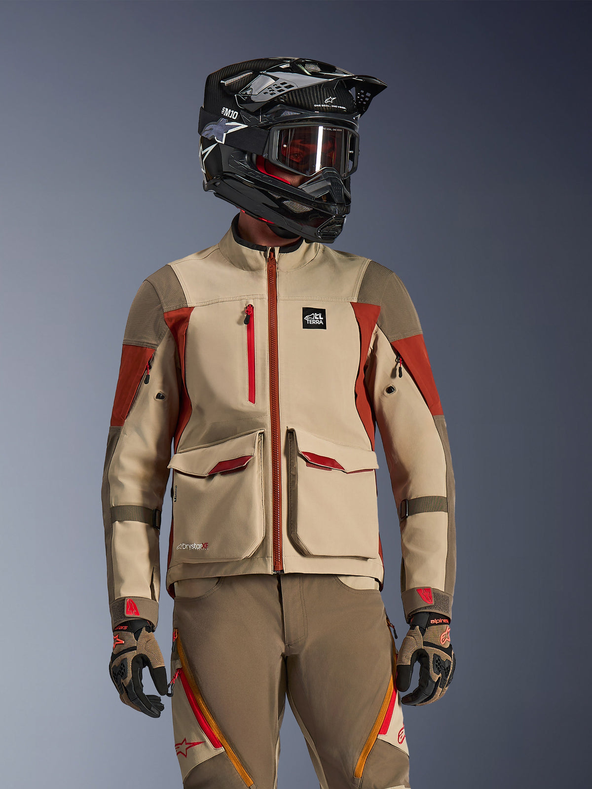 Adventure-Motorradfahrer trägt die Maxdura Drystar®XF Laminated Armored Shell in Aluminum Walnut Rust Brown, technische 3-Lagen-Konstruktion mit großen vorderen Cargo-Taschen und roten Akzenten, kombiniert mit einem schwarzen SM10-Helm, getönter Brille sowie passender Adventure-Hose und Handschuhen.