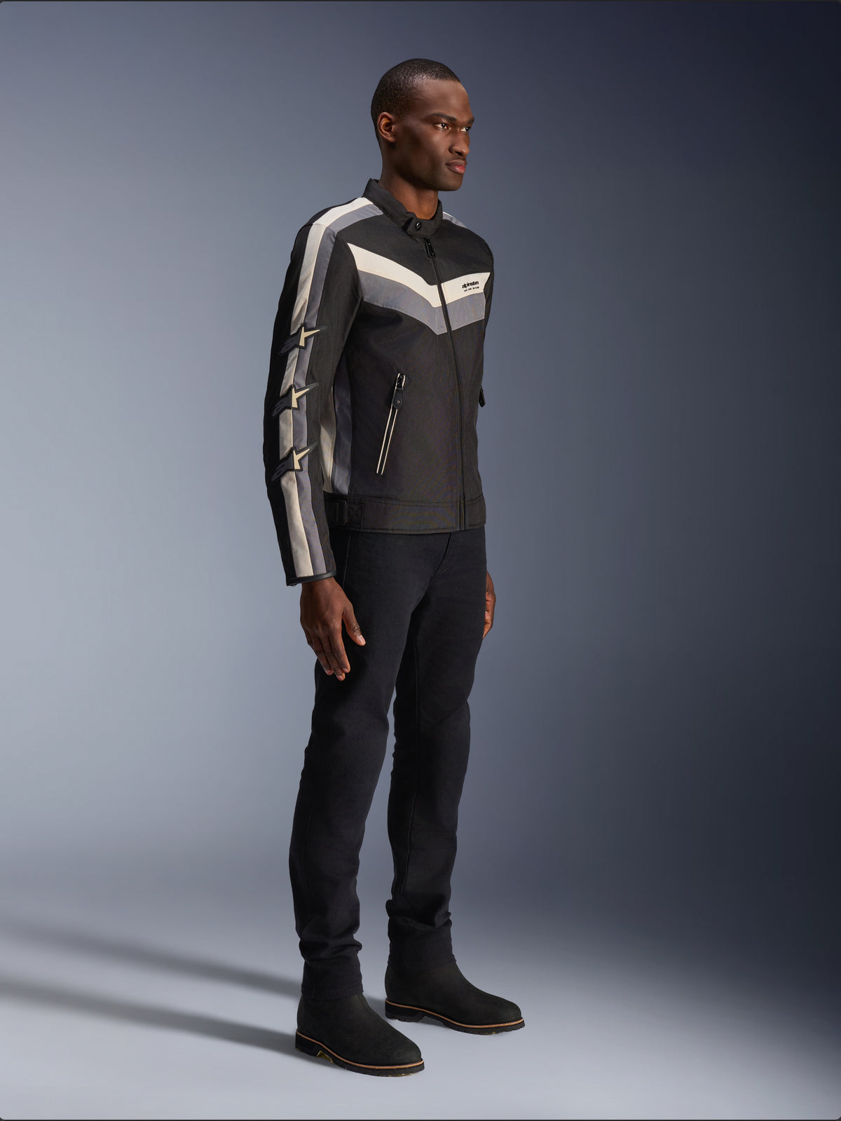 Alpinestars T-Dyno WR Jas, textiele motorjas, zwart met donkergrijze en ecru chevron-accenten, vintage-geïnspireerde bikerpasvorm, gedragen met zwarte broek en laarzen voor een complete road-riding look