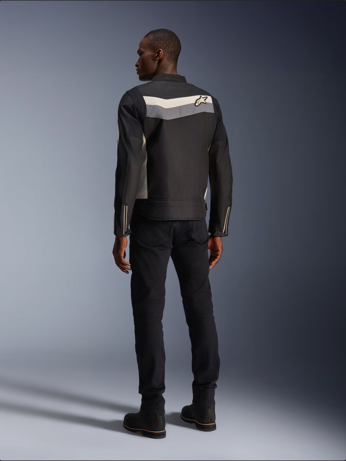 Mannelijk model van achteren gezien die de Alpinestars T-Dyno WR Jas in Black Dark Gray Ecru draagt, textiele motorjas met een horizontale streep en sterlogo op de rug, gecombineerd met een zwarte motorbroek en laarzen, urban motoruitrusting esthetiek.